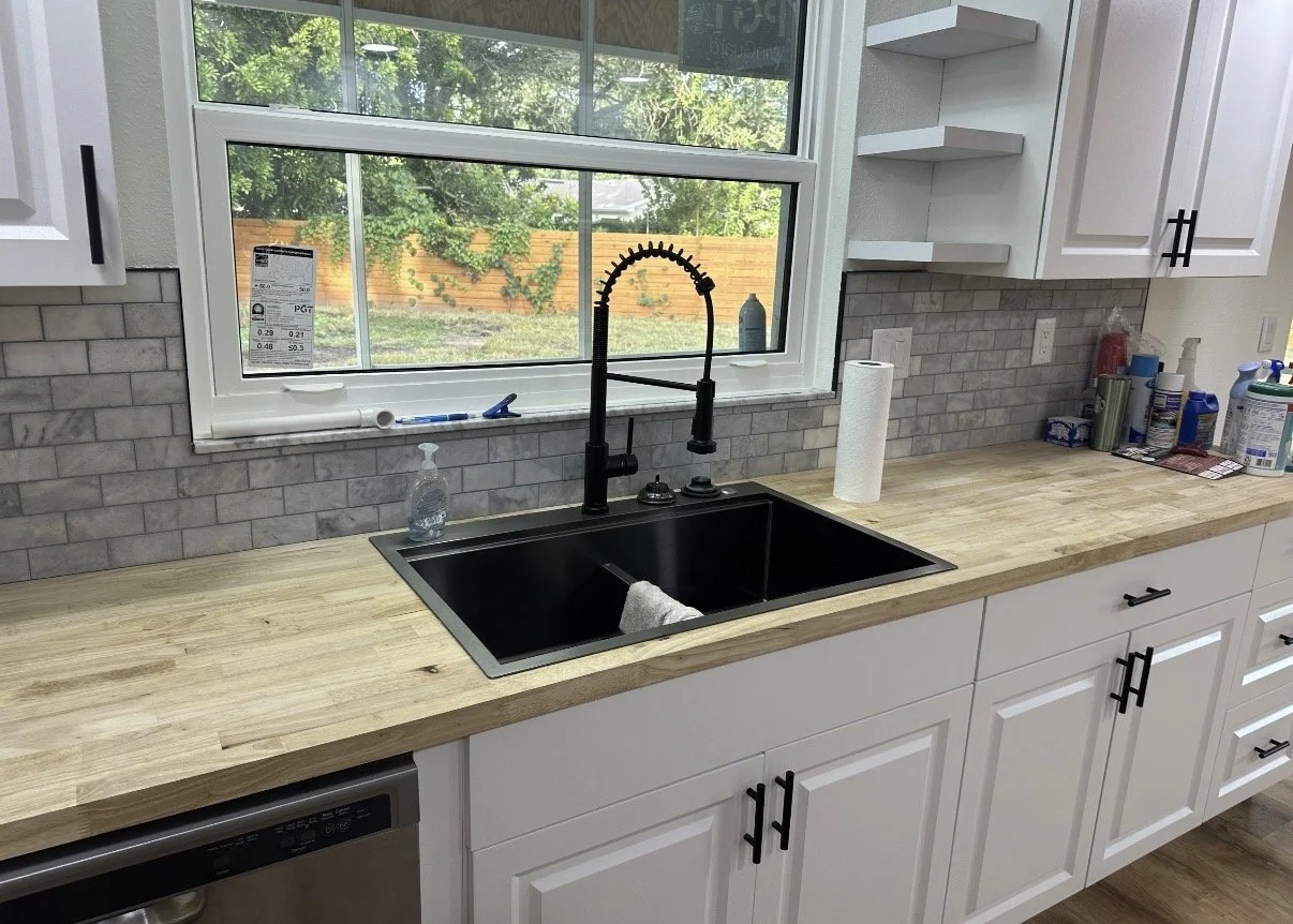 Kitchen countertop with a black sink, modern black faucet, paper towel roll, cleaning supplies, and a window showing a garden with trees and a wooden fence.