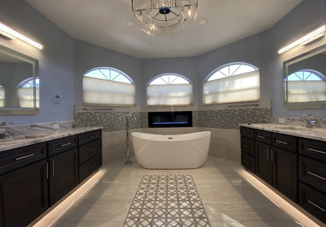 A luxurious bathroom featuring a freestanding bathtub between two double vanities with black cabinets and marble countertops. Four arched windows with beige shades, a modern chandelier, and decorative tile accents complete the space.