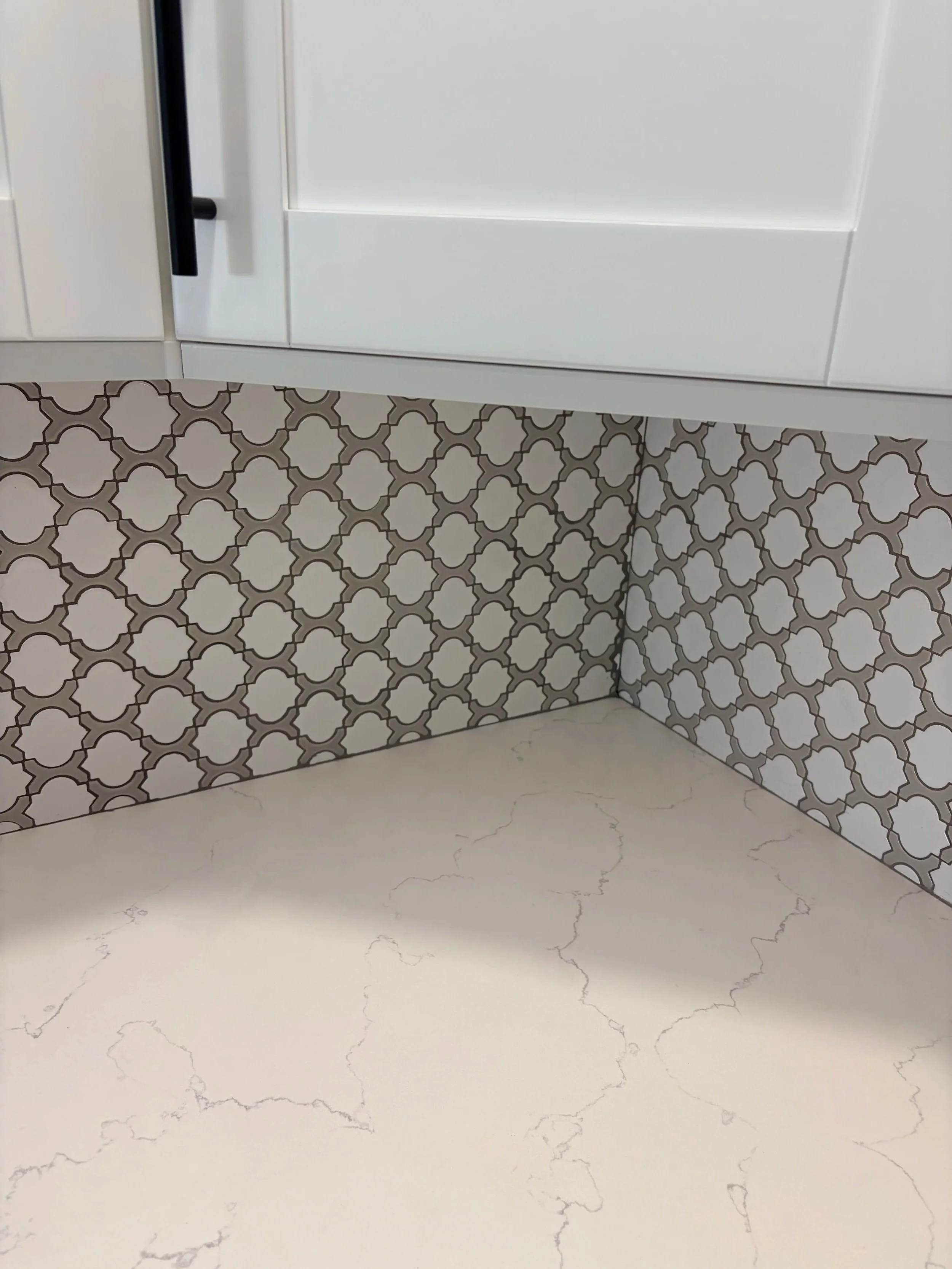 Close-up of a kitchen countertop with a white base and light gray veining, with a section of a wall covered in white and gray patterned tile.