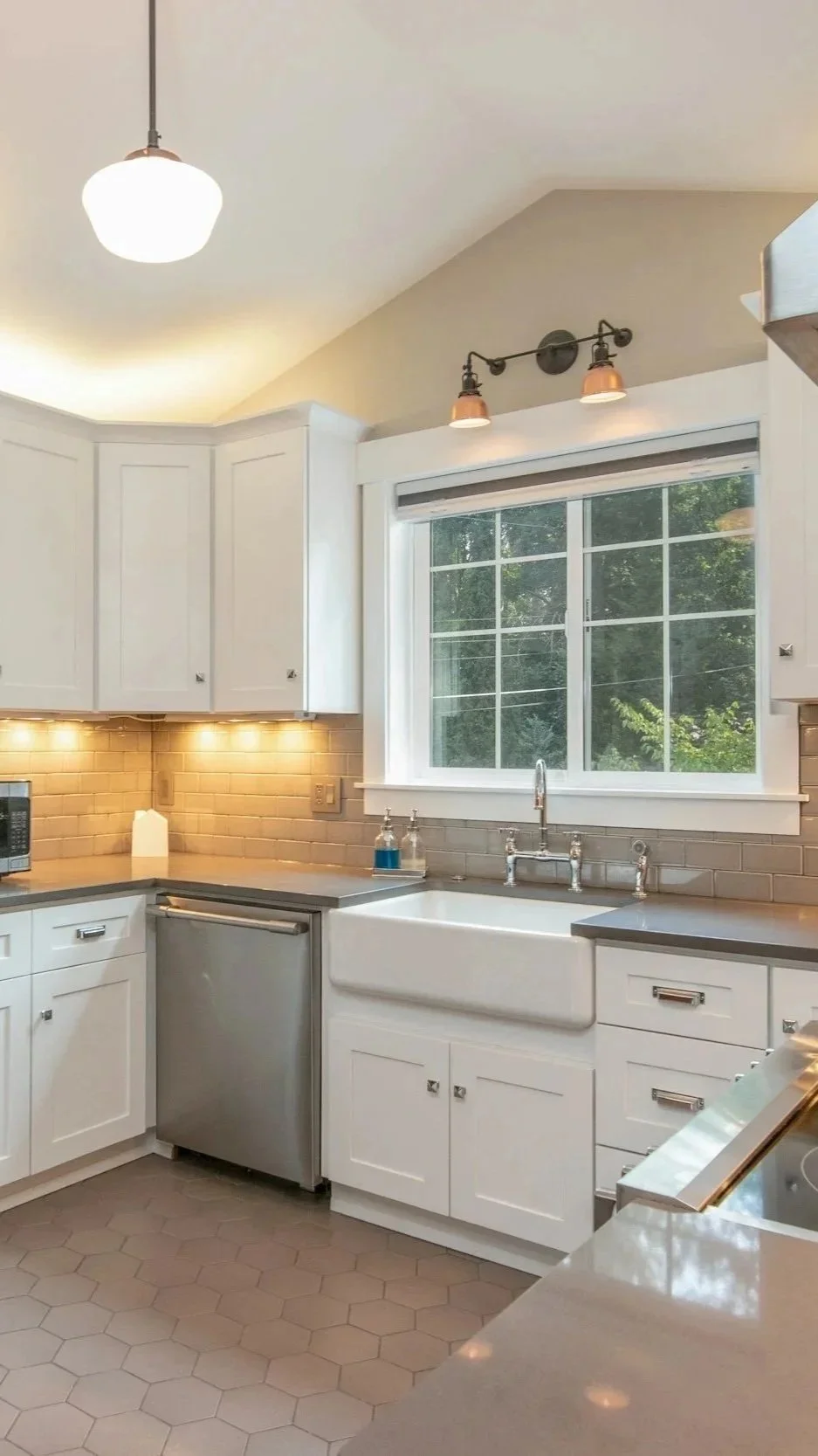 A modern kitchen with white cabinets, a large window, a farmhouse sink, and a beige tiled backsplash, illuminated by ceiling and under-cabinet lights.