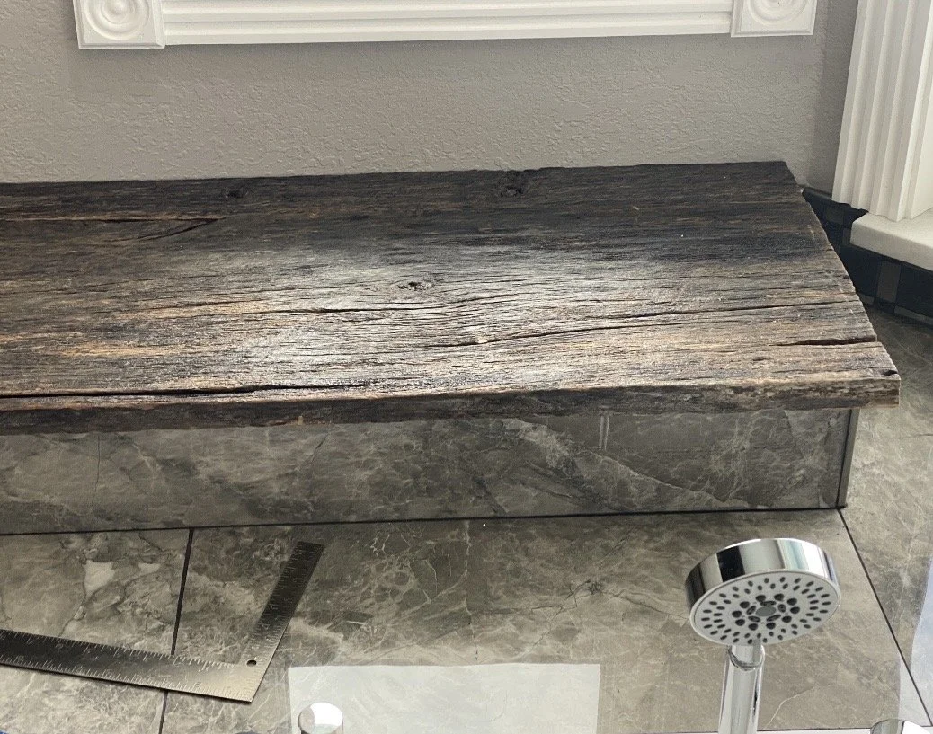 Close-up of a rustic wooden bench with a dark, weathered finish, placed on a gray stone tiled floor near a gray wall.