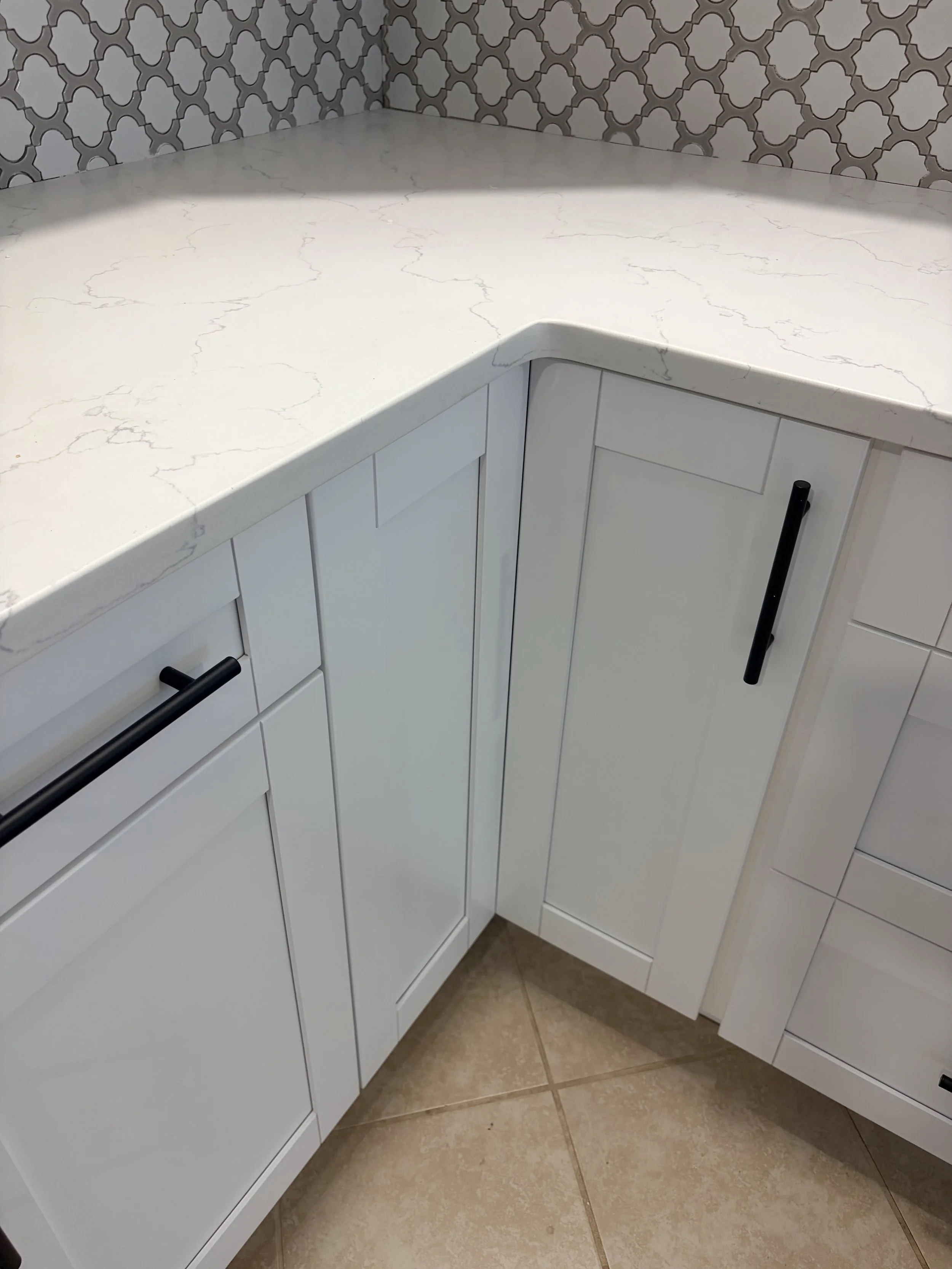 White kitchen cabinets with black handles, white marble countertops, tiled brown floor, and patterned wallpaper.