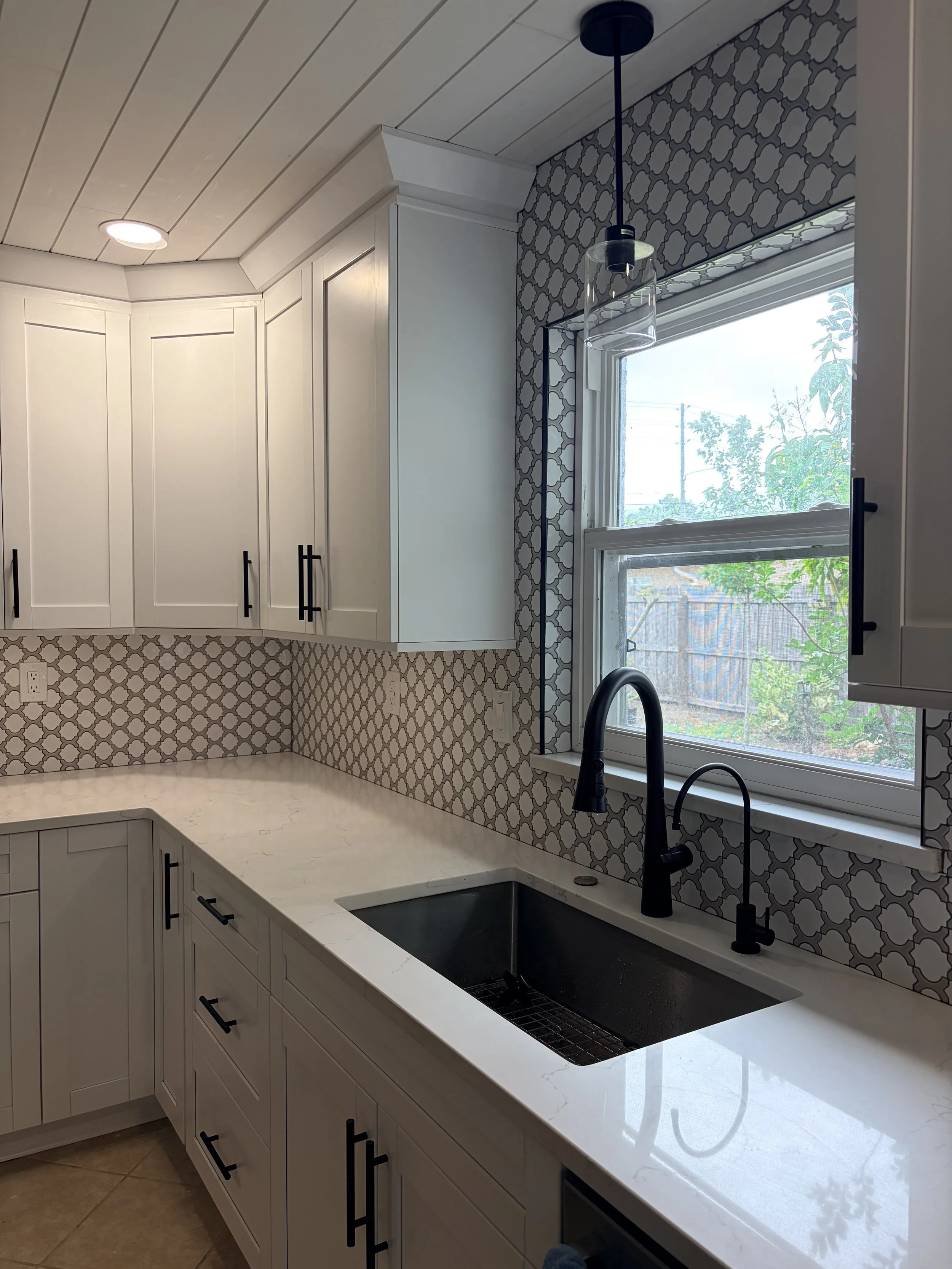 Kitchen interior featuring a white countertop, black sink and faucet, white cabinets with black handles, a window with a partly cloudy view outside, patterned backsplash and ceiling, and pendant light hanging from the ceiling.