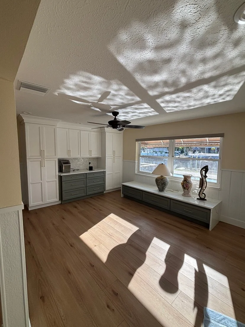 Empty living room with light wood flooring, white cabinets, a ceiling fan, a window with outdoor view, and decorative vases and a lamp on a low storage bench.