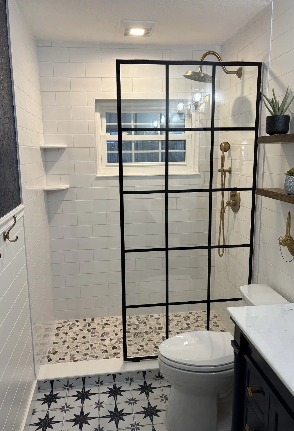 Modern bathroom with black-framed glass shower enclosure, white subway tile walls, pebble stone shower floor, window, gold fixtures, patterned floor tiles, and decorative shelving with plants.