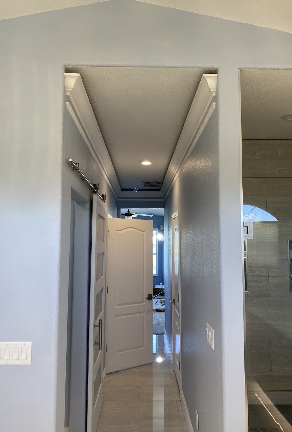 Photo of a home hallway with an open door at the end, multiple doors along the hallway, light-colored walls, and a ceiling with crown molding and recessed lighting.