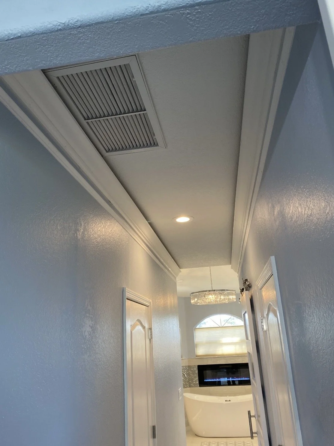 Hallway leading to a bathroom with a whirlpool bathtub, a window with blinds, a chandelier, and a dark wall-mounted TV.