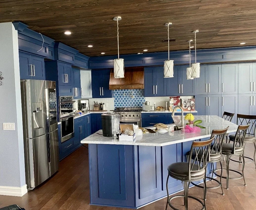 Blue kitchen with white marble island, four pendant lights, stainless steel refrigerator, oven, coffee machine, stove with blue tiled backsplash, and brown wooden ceiling with recessed lighting. Four bar stools around the island.