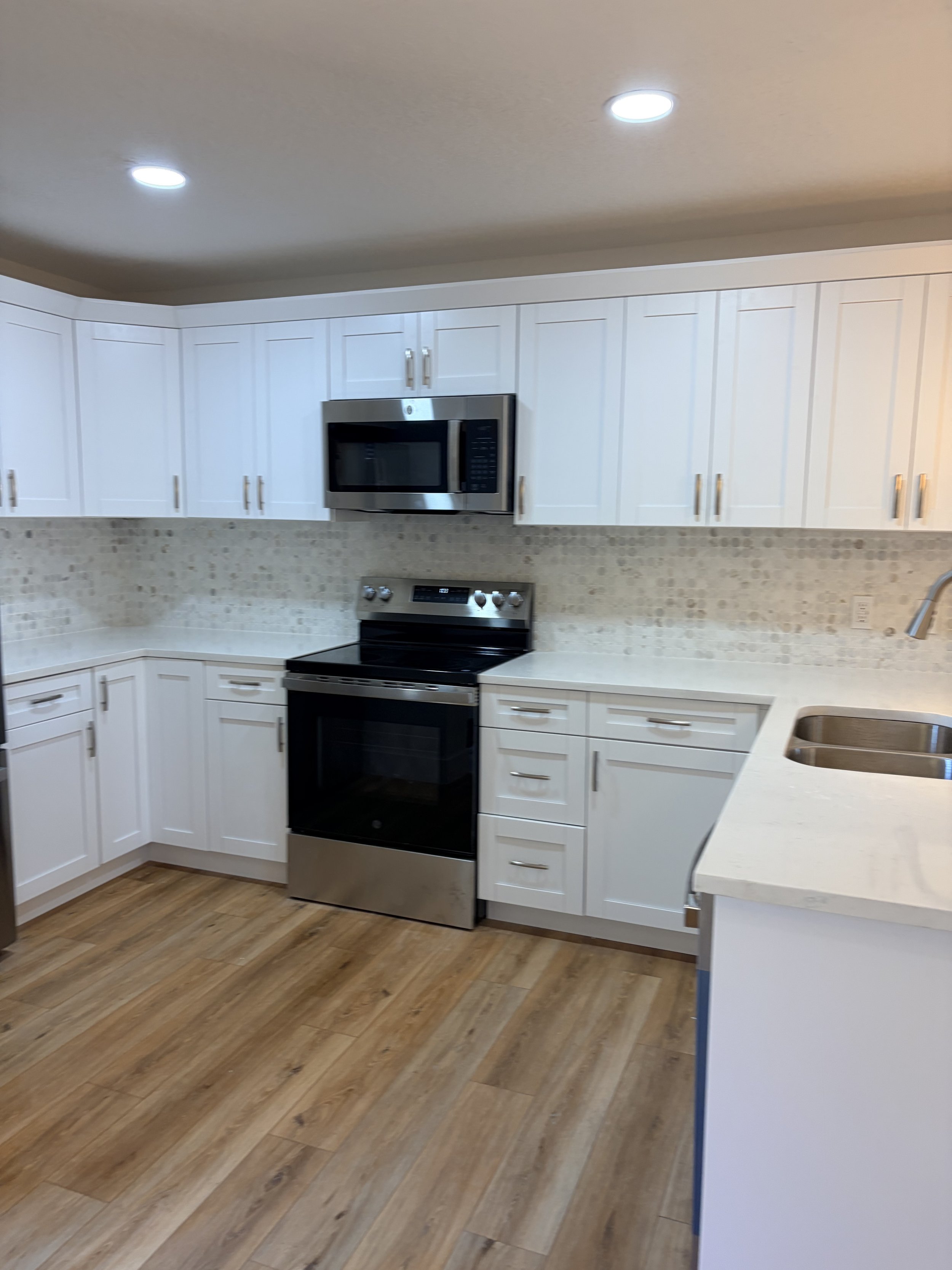 Modern kitchen with white cabinets, stainless steel microwave and oven, beige tile backsplash, and wooden flooring.