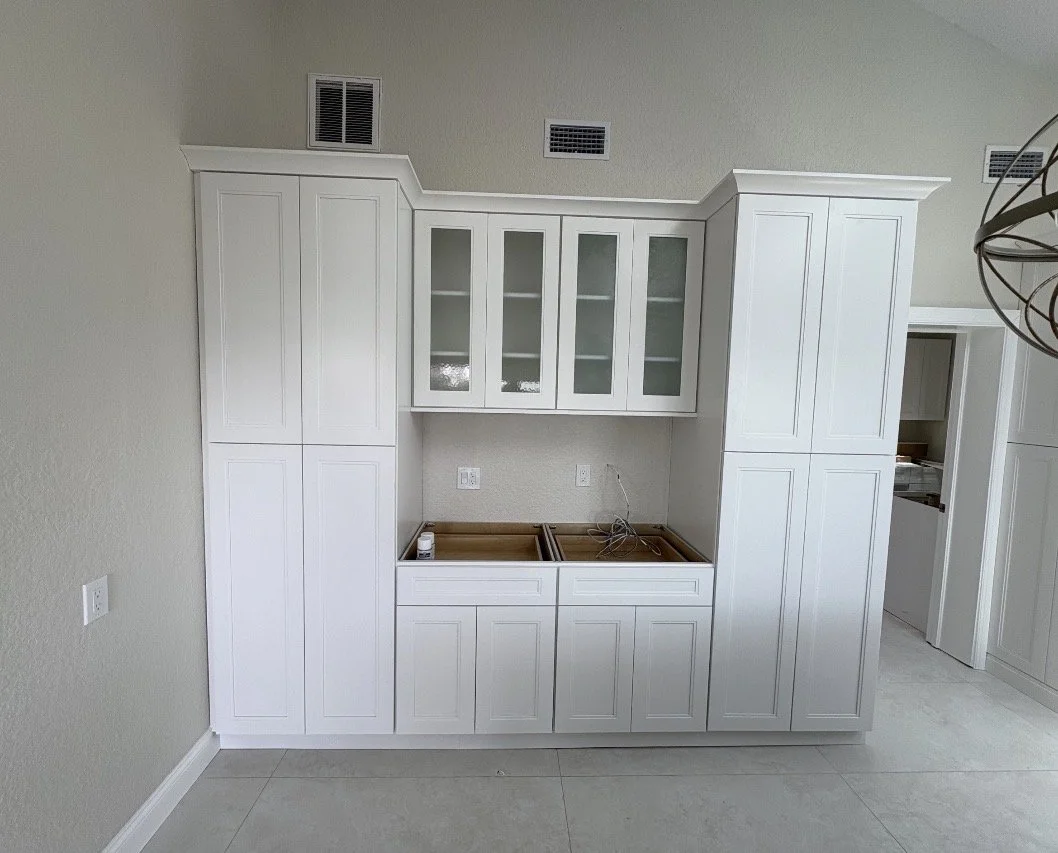 White kitchen cabinets with glass doors are being installed in a room, with some wiring visible and no countertop yet.