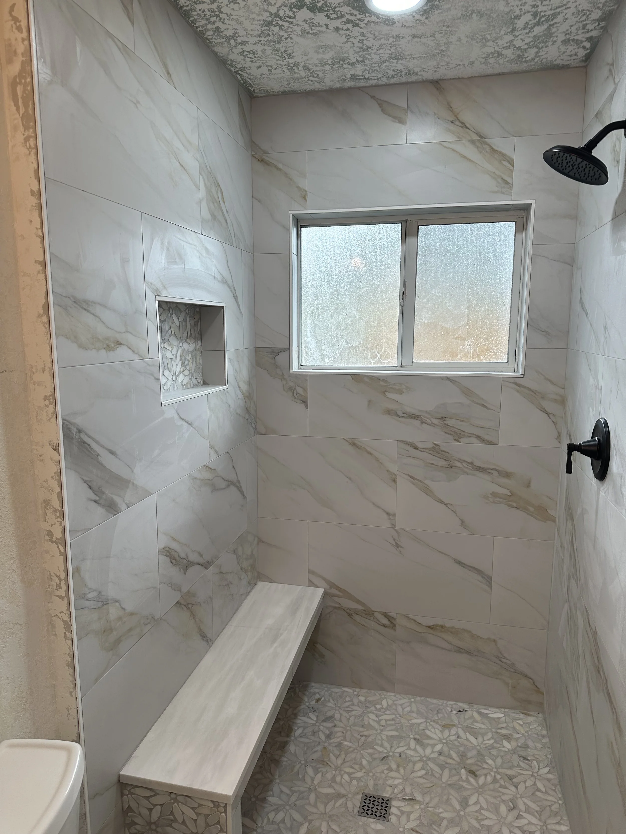 A modern shower with large white marble tiles on the walls, a built-in niche filled with small white stones, a frosted glass window, a black showerhead, and a white bench with a marble top. The floor has decorative pebble tiles with floral patterns and a square drain.