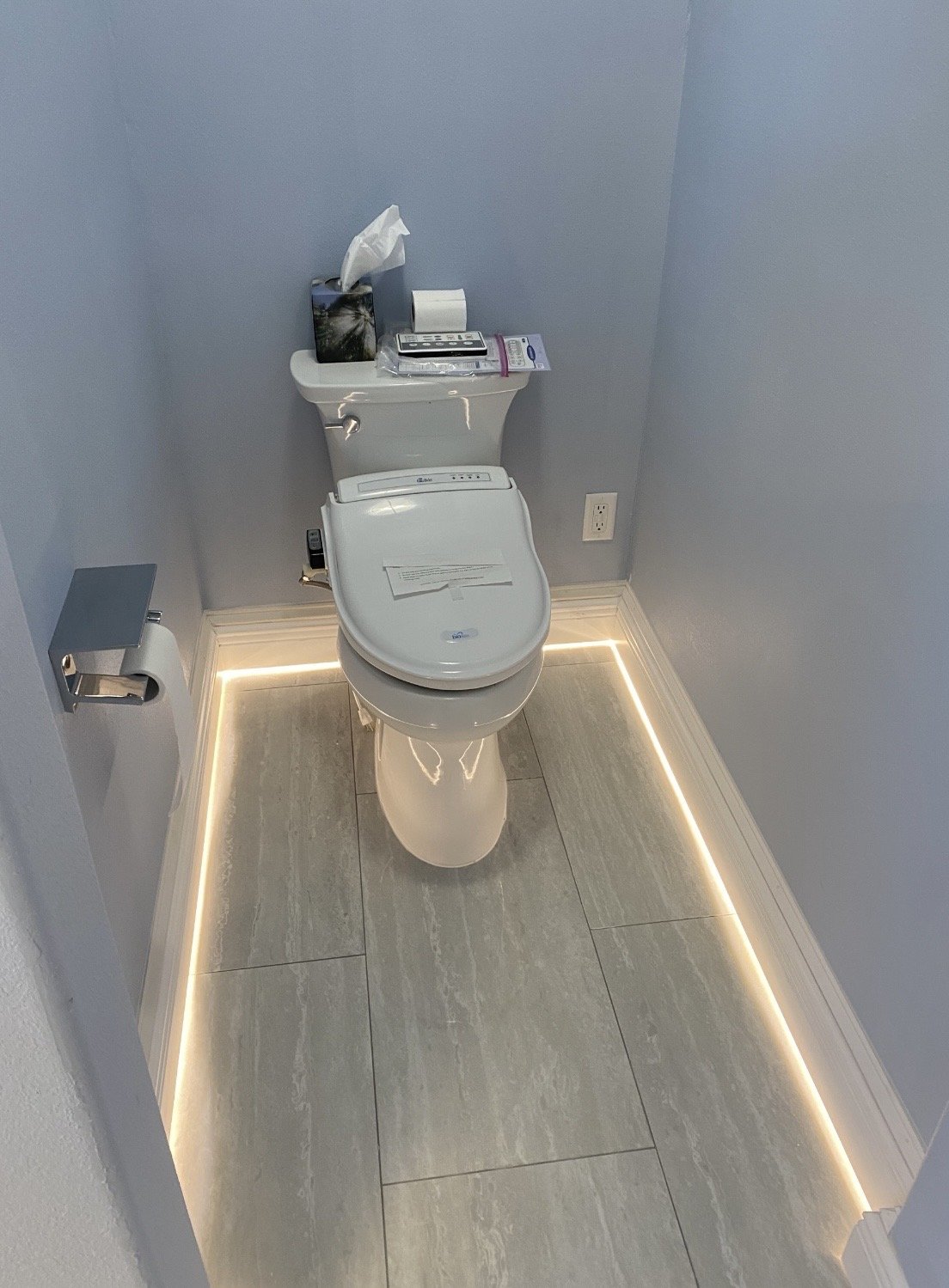 Modern toilet with a shelf on top holding tissue, electronics, and a package, in a small bathroom with LED lighting around the baseboards.