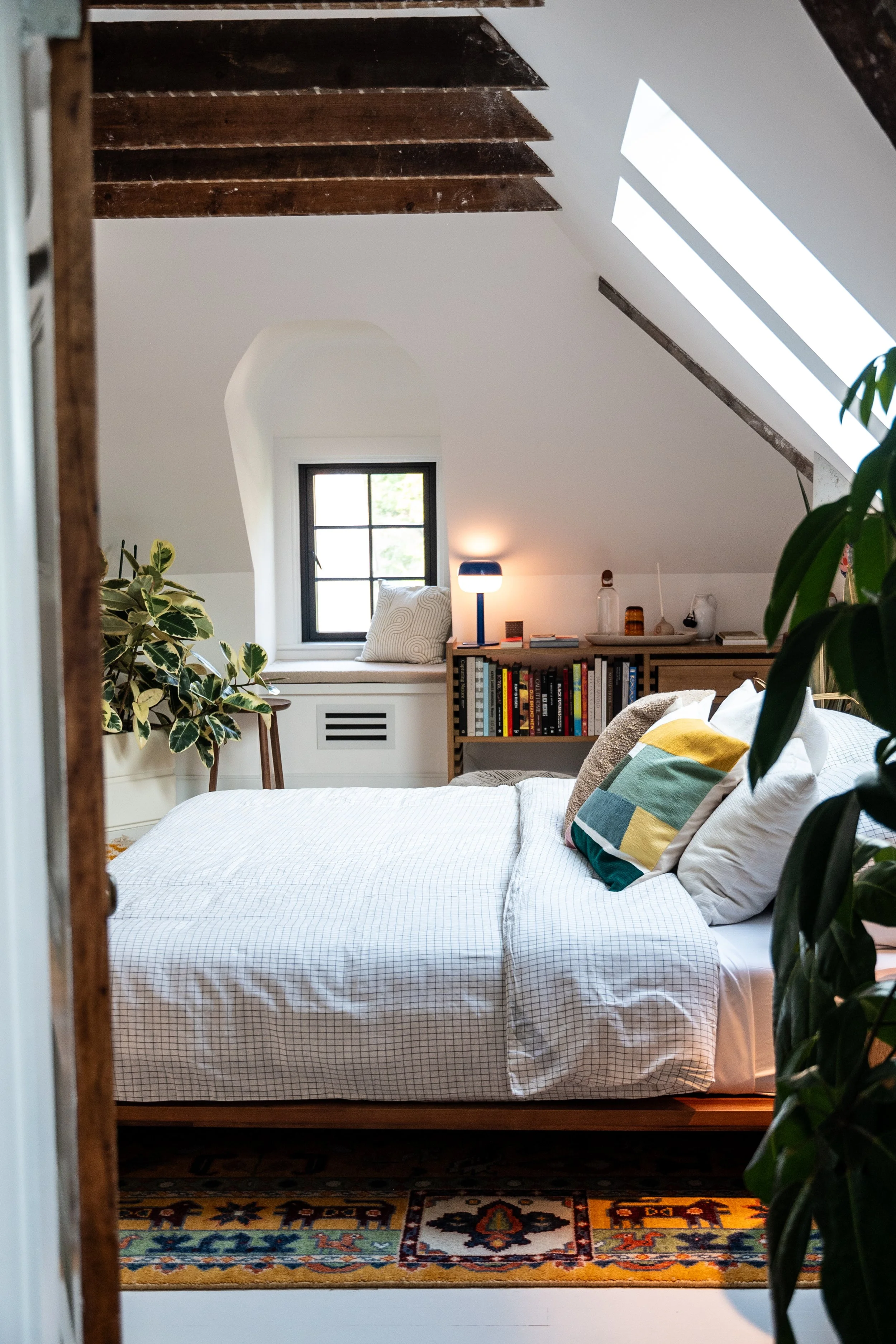 A cozy attic bedroom with a white bed, colorful pillows, a window seat with a cushion, a small bookshelf with books, a table lamp, and skylights in the ceiling.