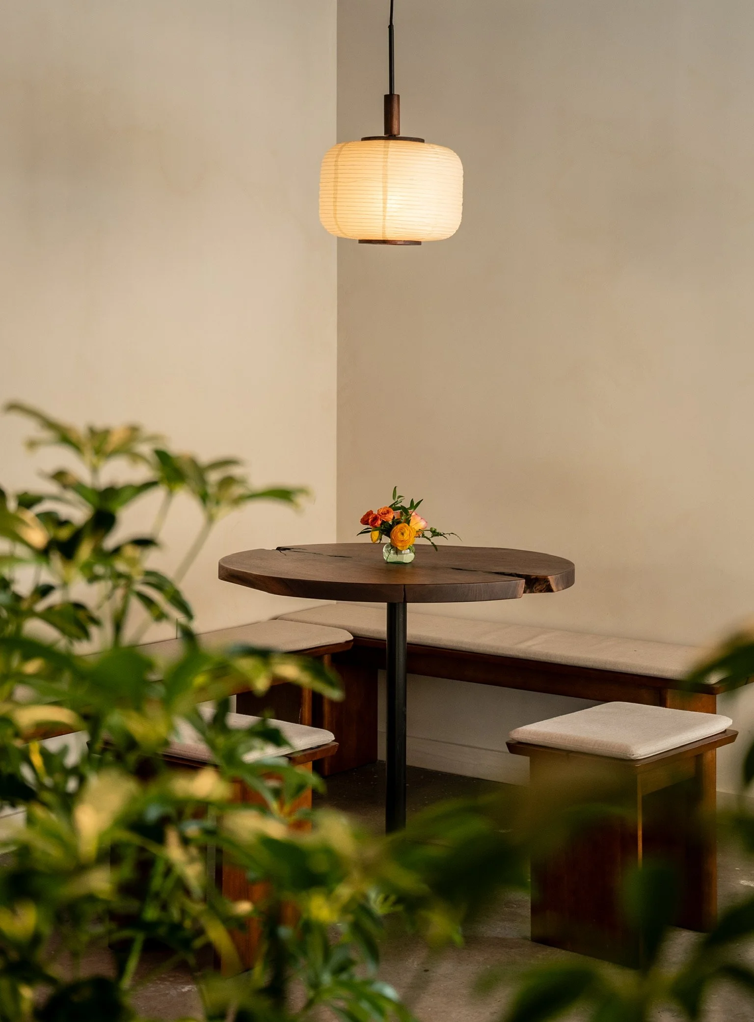 Interior view of a cozy dining area with a round wooden table, a floral centerpiece, and a hanging paper lantern light, surrounded by built-in seating with cushions, partially occluded by green plants in the foreground.