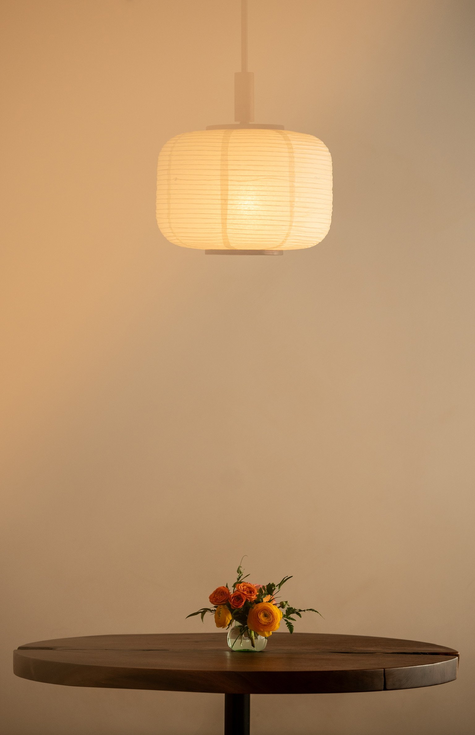 A wooden round table with a glass vase of orange and yellow flowers, illuminated by a beige paper lantern ceiling light against a plain beige wall.