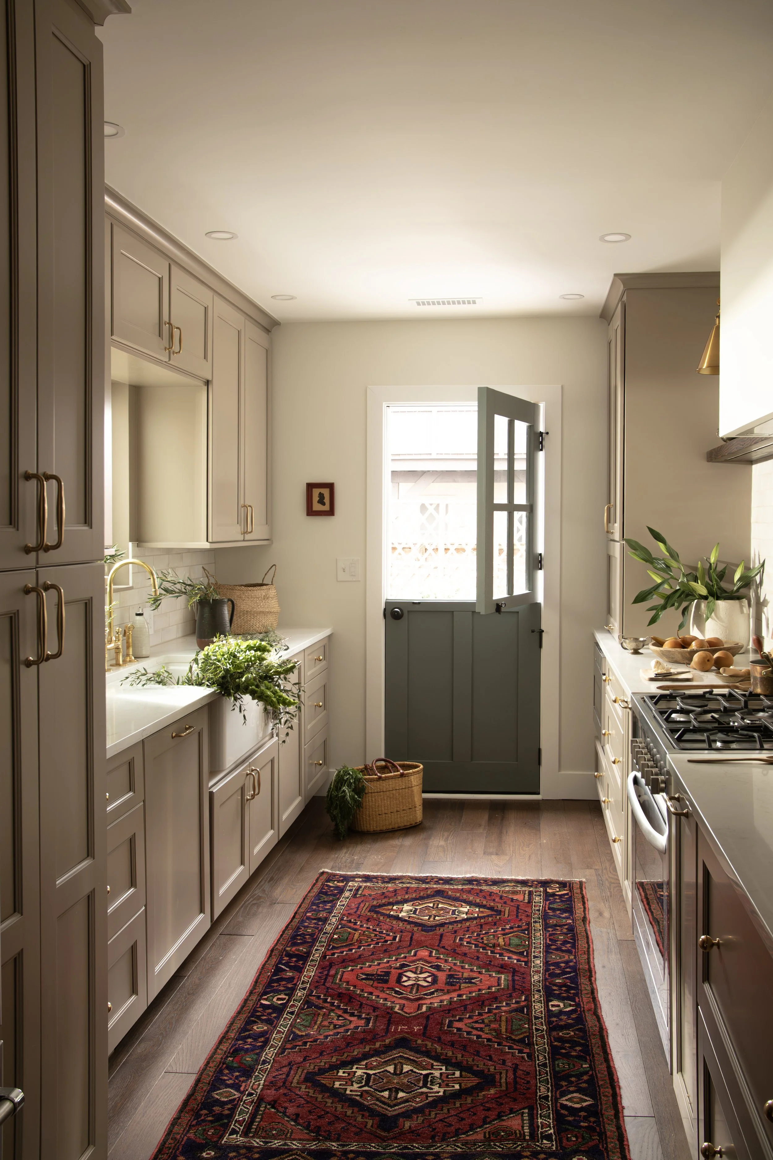 A cozy kitchen with light gray cabinets, wooden flooring, and a colorful patterned rug in the center. Natural light streams in through a window and a partially open door at the back, revealing outdoor space. Green plants and a woven basket with green