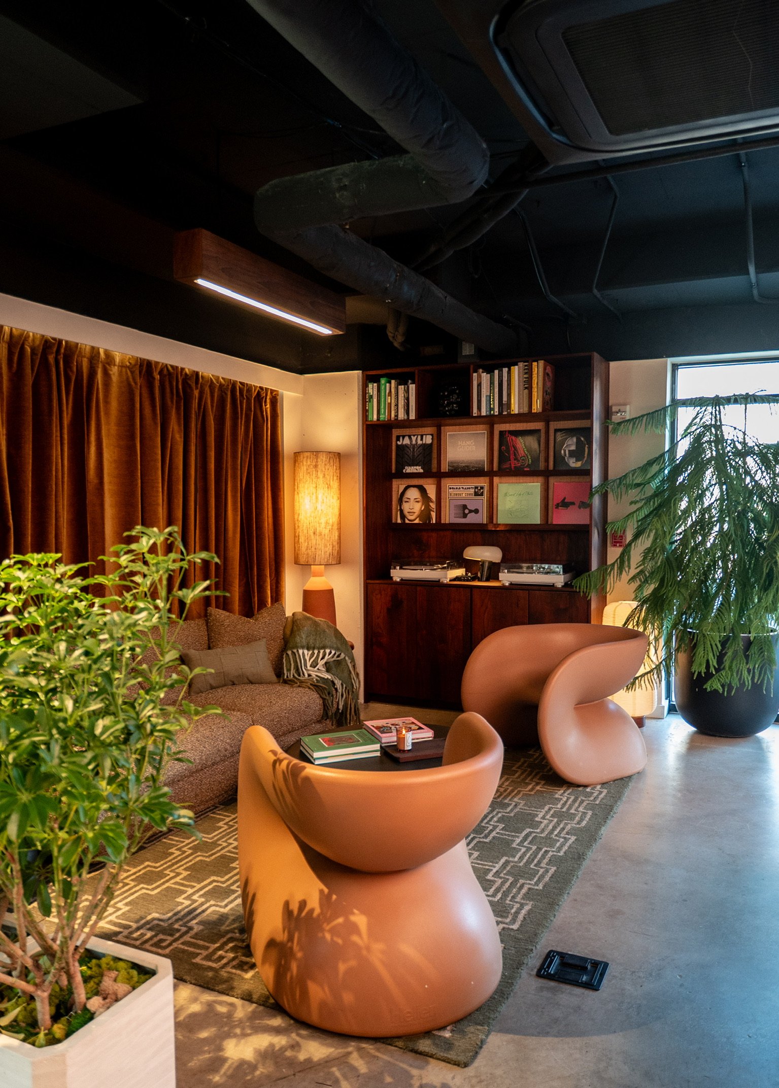 A cozy living room with a brown sofa, pink modern chairs, a wooden bookshelf with records and books, a tall lamp, green plants, a large window, a patterned rug, and industrial ceiling pipes.