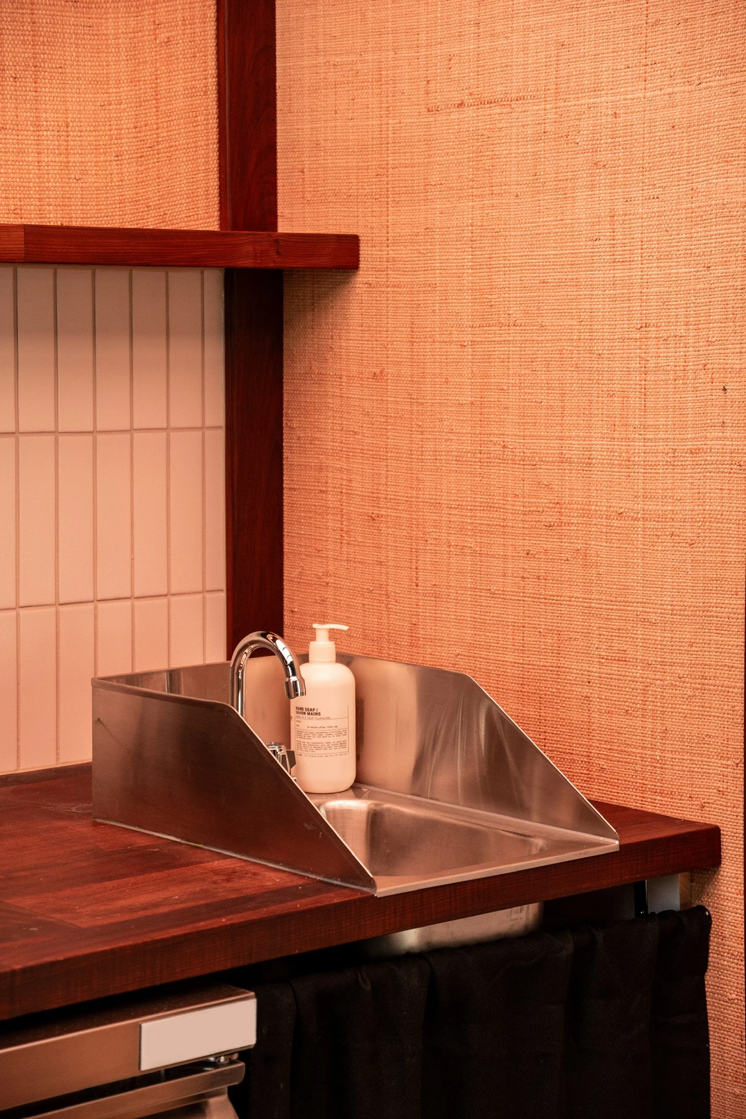 A stainless steel sink with a soap dispenser on the side, set on a wooden countertop. The background has a textured orange wall and part of a tiled wall with white tiles.