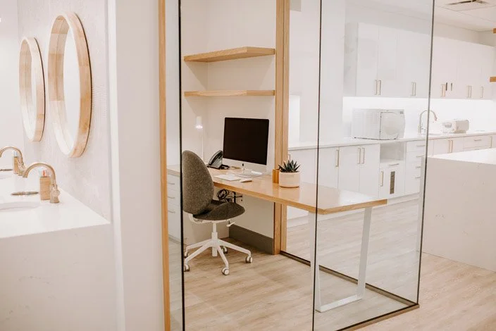 Modern home office space with a desk, computer, chair, and floating shelves, separated by glass walls, adjacent to a kitchen area.