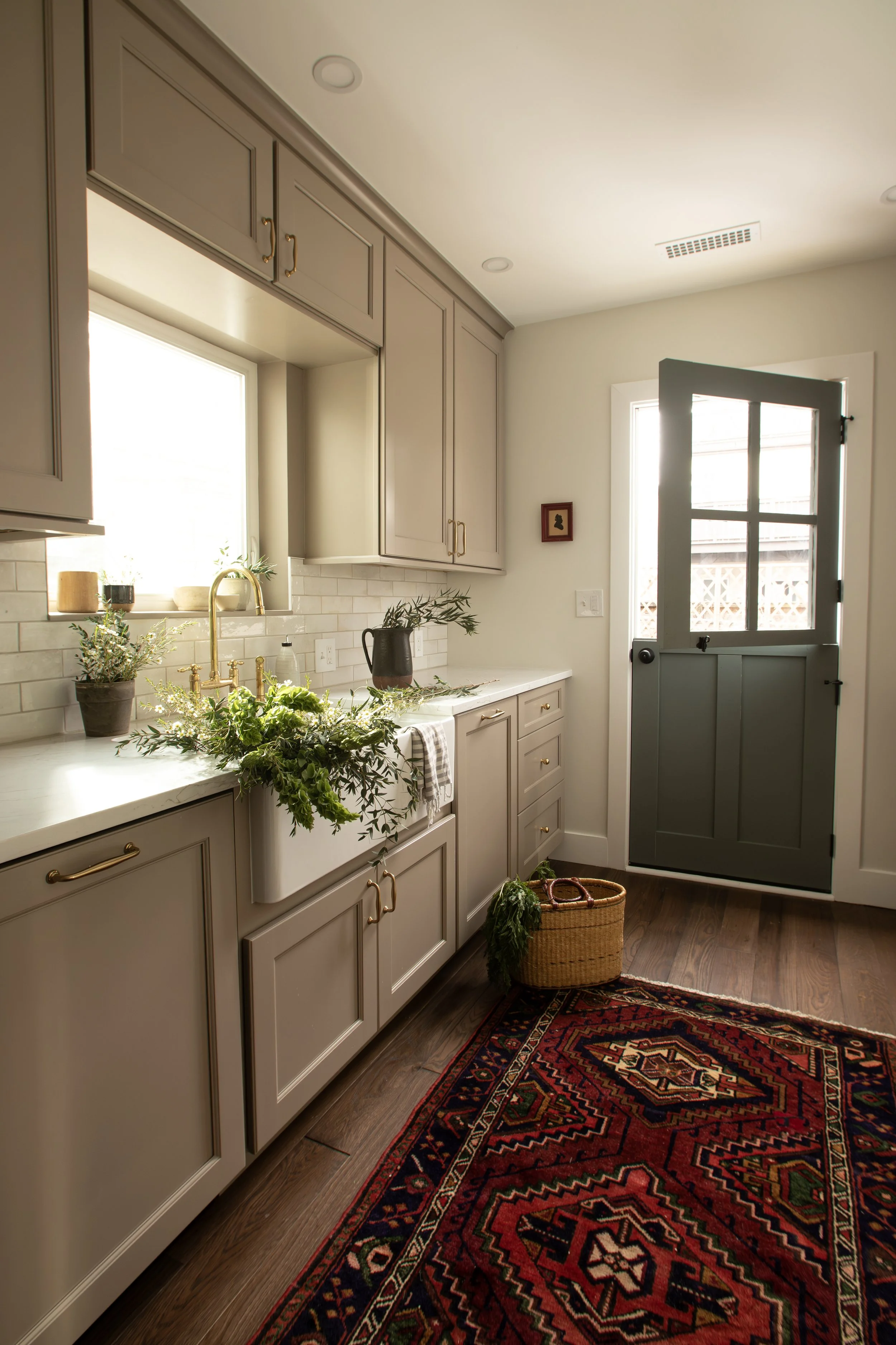 A cozy kitchen with beige cabinets, a white farmhouse sink, a floral arrangement, and a dark green door with window panes. A colorful patterned rug and a basket with greenery are on the wooden floor.