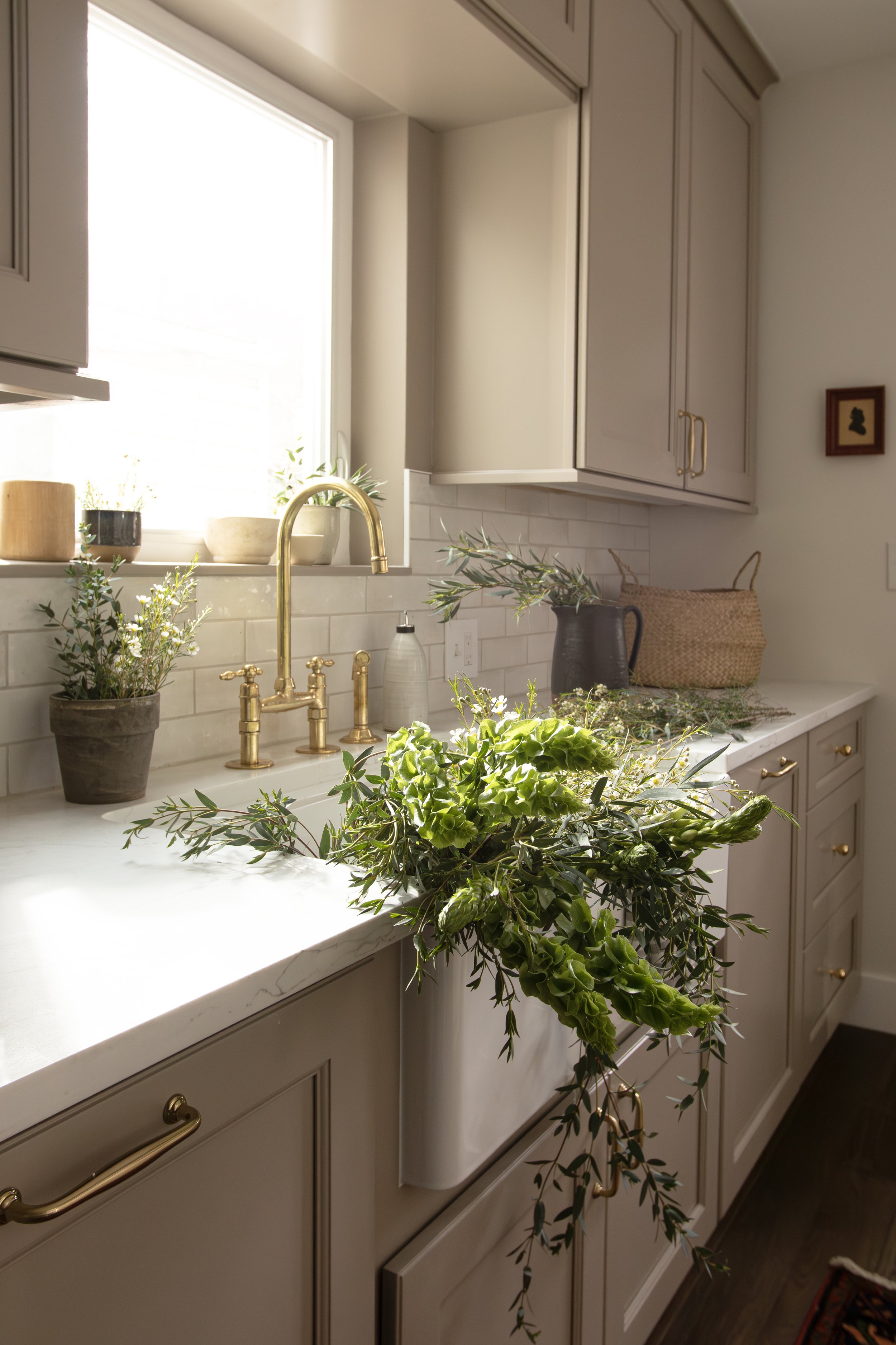 Kitchen countertop with a window above, decorated with plants, a soap dispenser, and a basket, featuring a fresh floral arrangement.