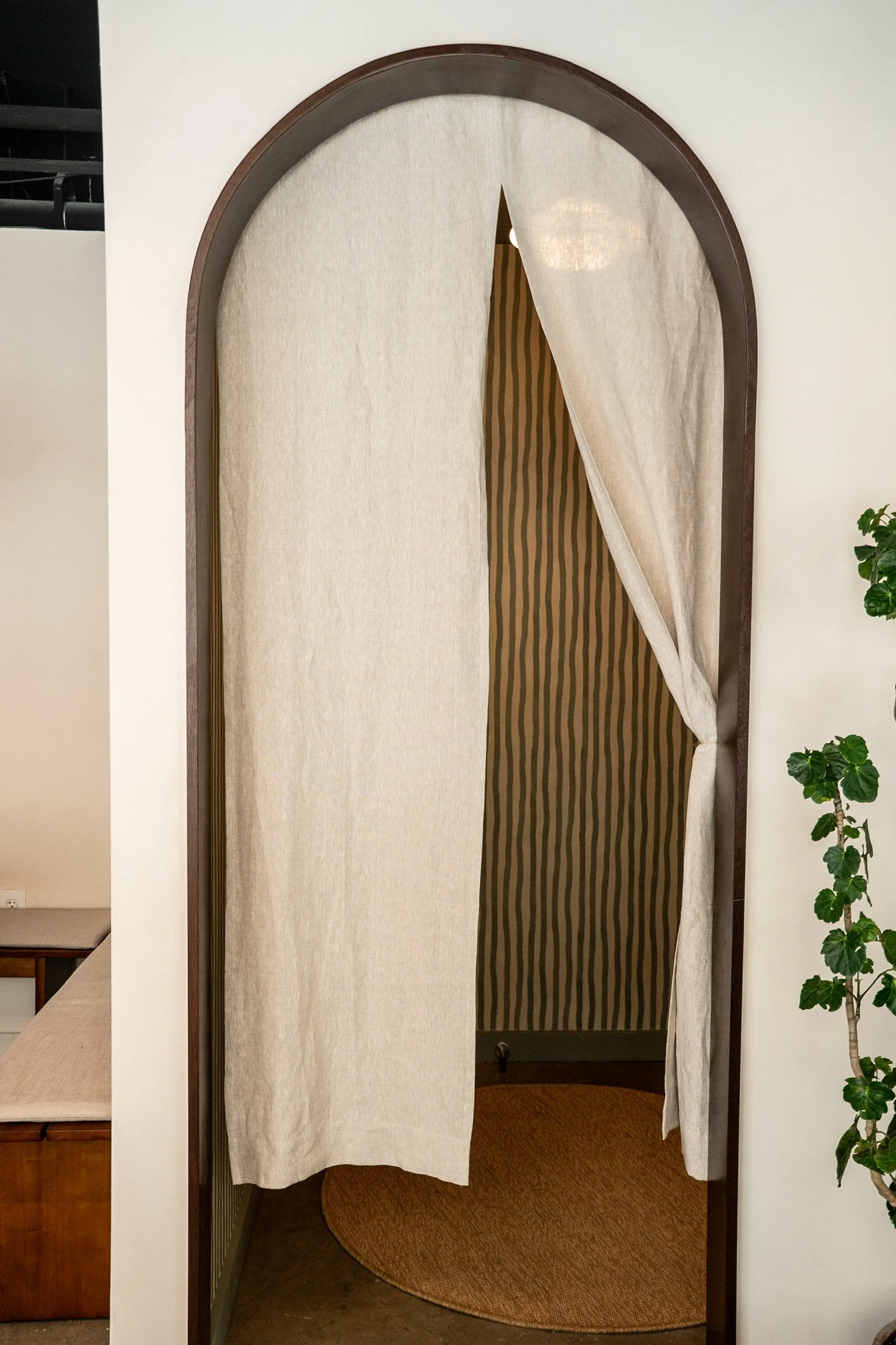 A fitting room with beige curtains, one pulled back, inside a store. A round brown rug is on the floor.