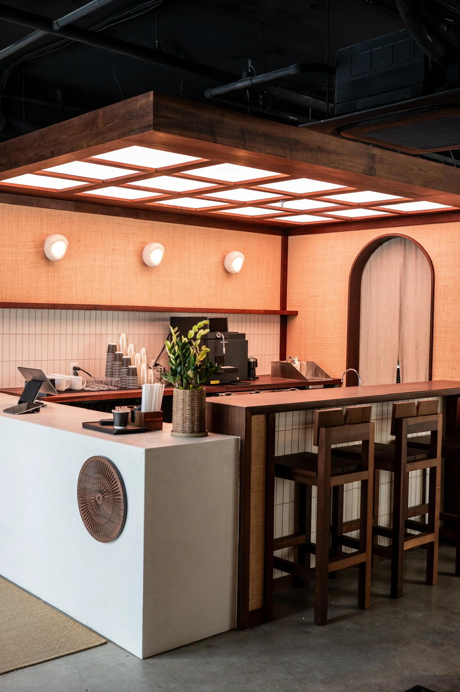Interior of a modern coffee bar with wooden counters, bar stools, a plant, and a white counter with cups, glasses, and coffee supplies.