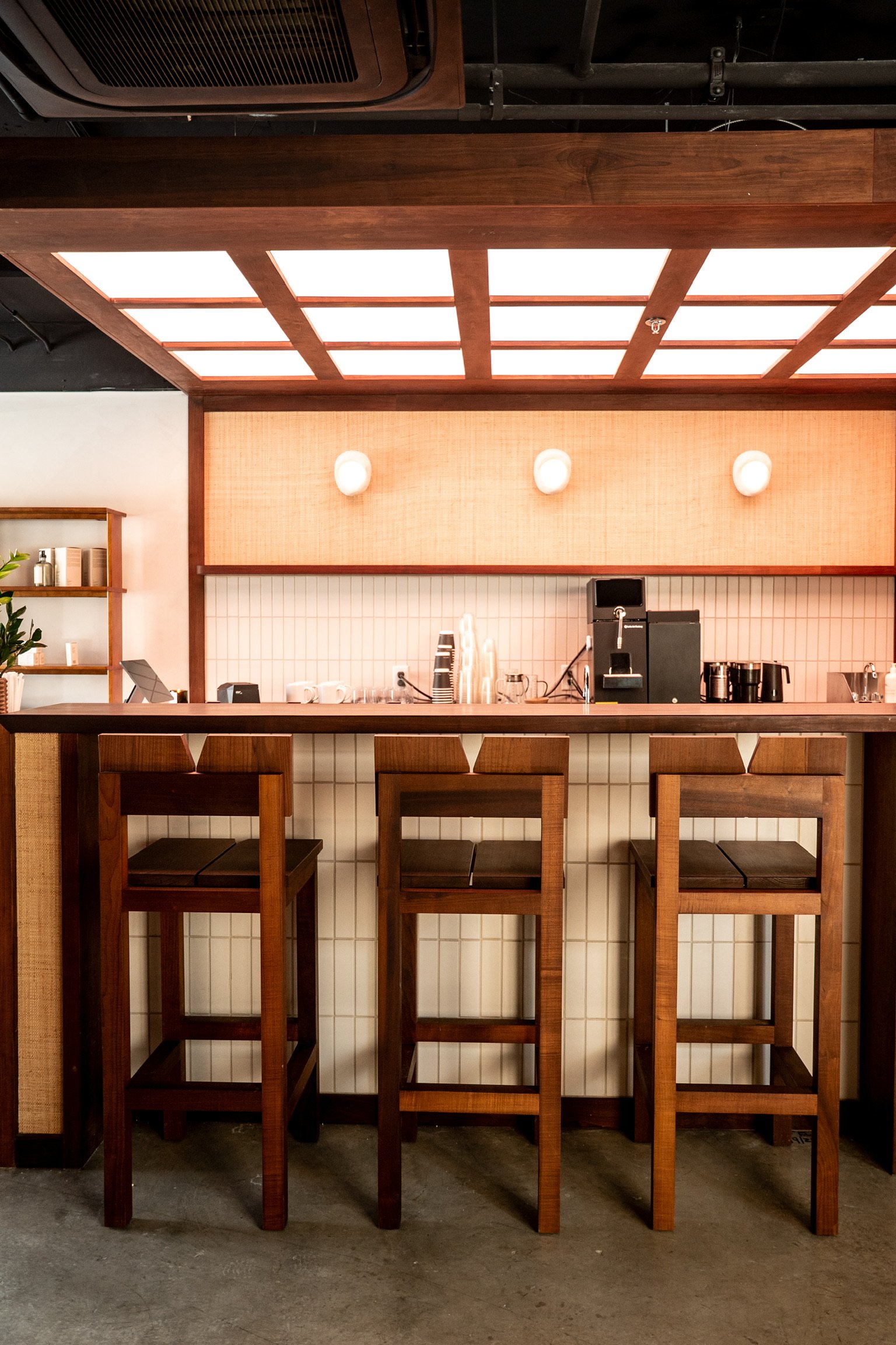 View of a modern cafe or restaurant counter area with a wooden bar, three tall wooden chairs, a coffee machine, cups, and a light wood accent wall. The ceiling has a wooden grid design with illuminated panels.