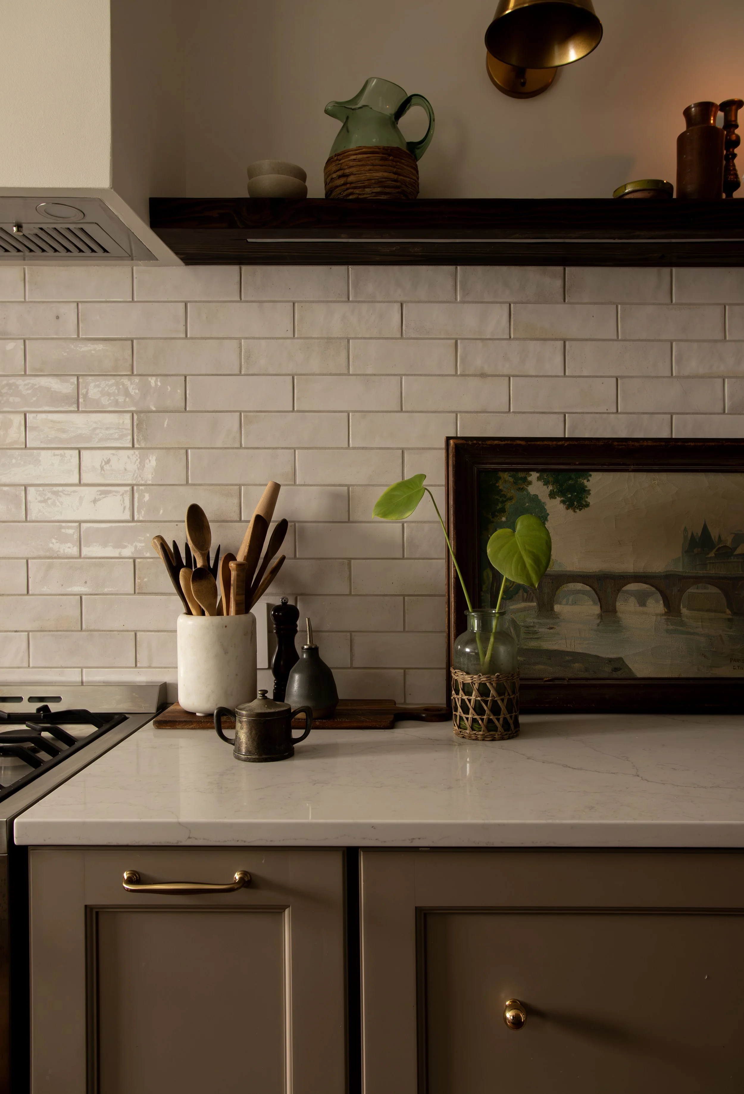 Interior view of a kitchen counter with a container of wooden cooking utensils, a framed landscape painting, a green potted plant, and various ceramic and metal decor items on a countertop with a white brick backsplash and dark cabinetry.