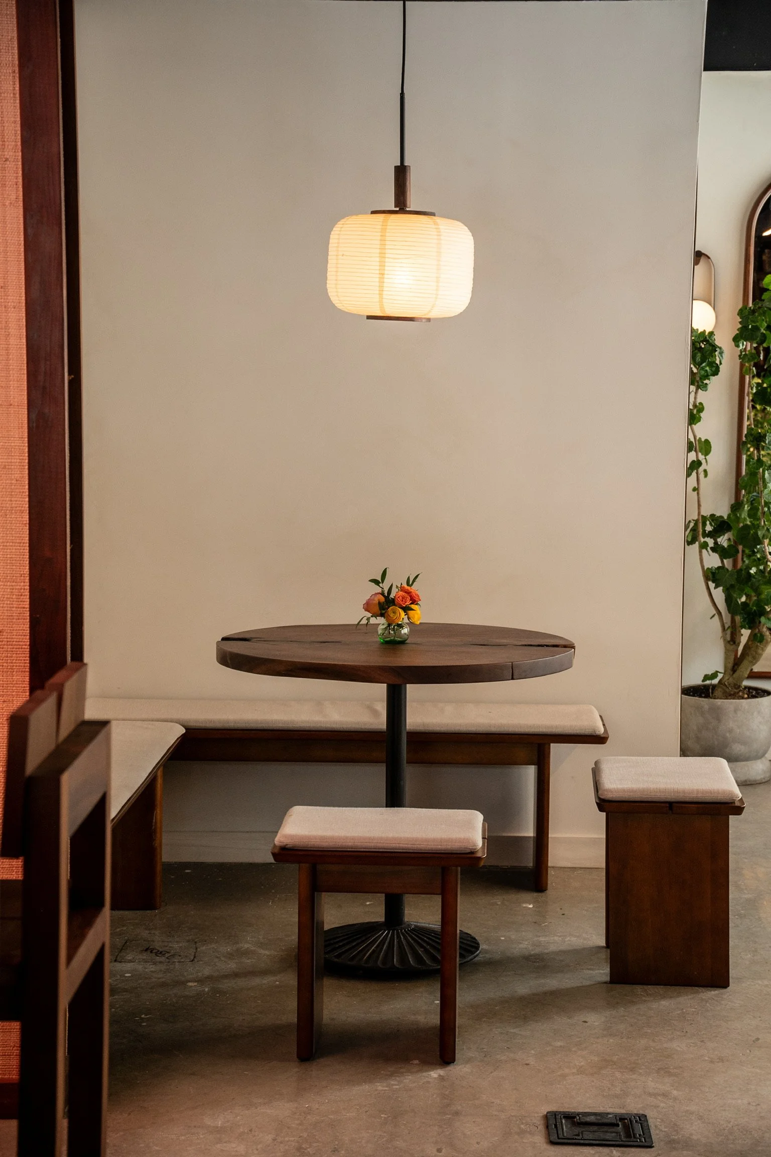 A cozy corner in a cafe with a round wooden table, cushioned stools, a small vase of orange and yellow roses, a hanging paper lantern, and greenery on the right side.