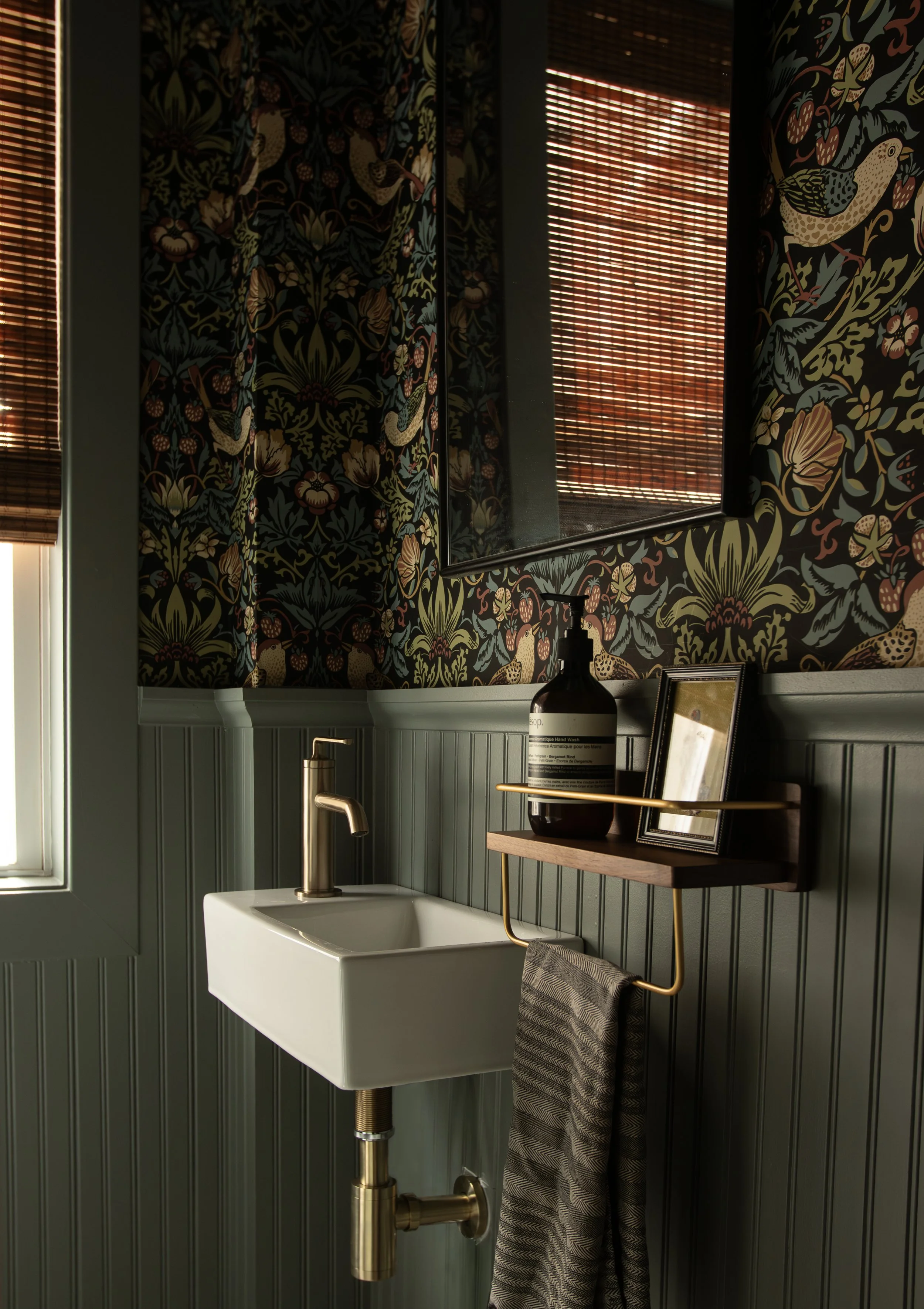 Small bathroom with a white wall-mounted sink, bronze faucet, and a black shelf holding a large black soap dispenser, a framed mirror, and a black and white striped towel hanging from a gold towel bar. The upper part of the walls has dark floral wallpaper, and there is a window with bamboo blinds.