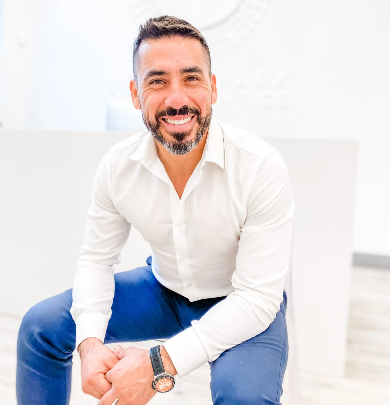 A smiling man with dark hair, a beard, and mustache, wearing a white dress shirt and blue pants, sitting in a bright, modern office space.