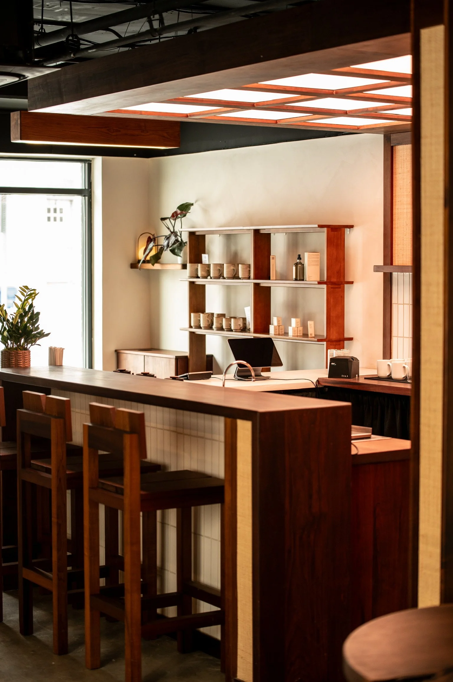 Interior of a modern cafe or restaurant with wooden furniture, shelves with mugs and decorative plants, and a large window letting in natural light.