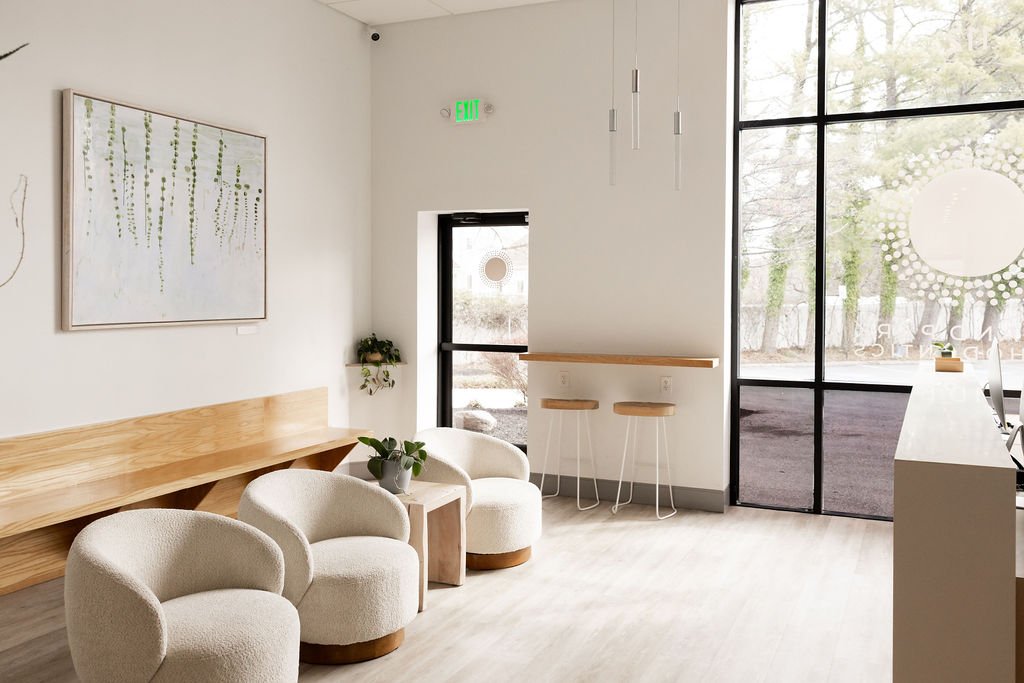 A modern, minimalistic waiting area with white walls, a wooden bench, three white rounded armchairs, a small wooden side table with a potted plant, and a large window showing trees outside.