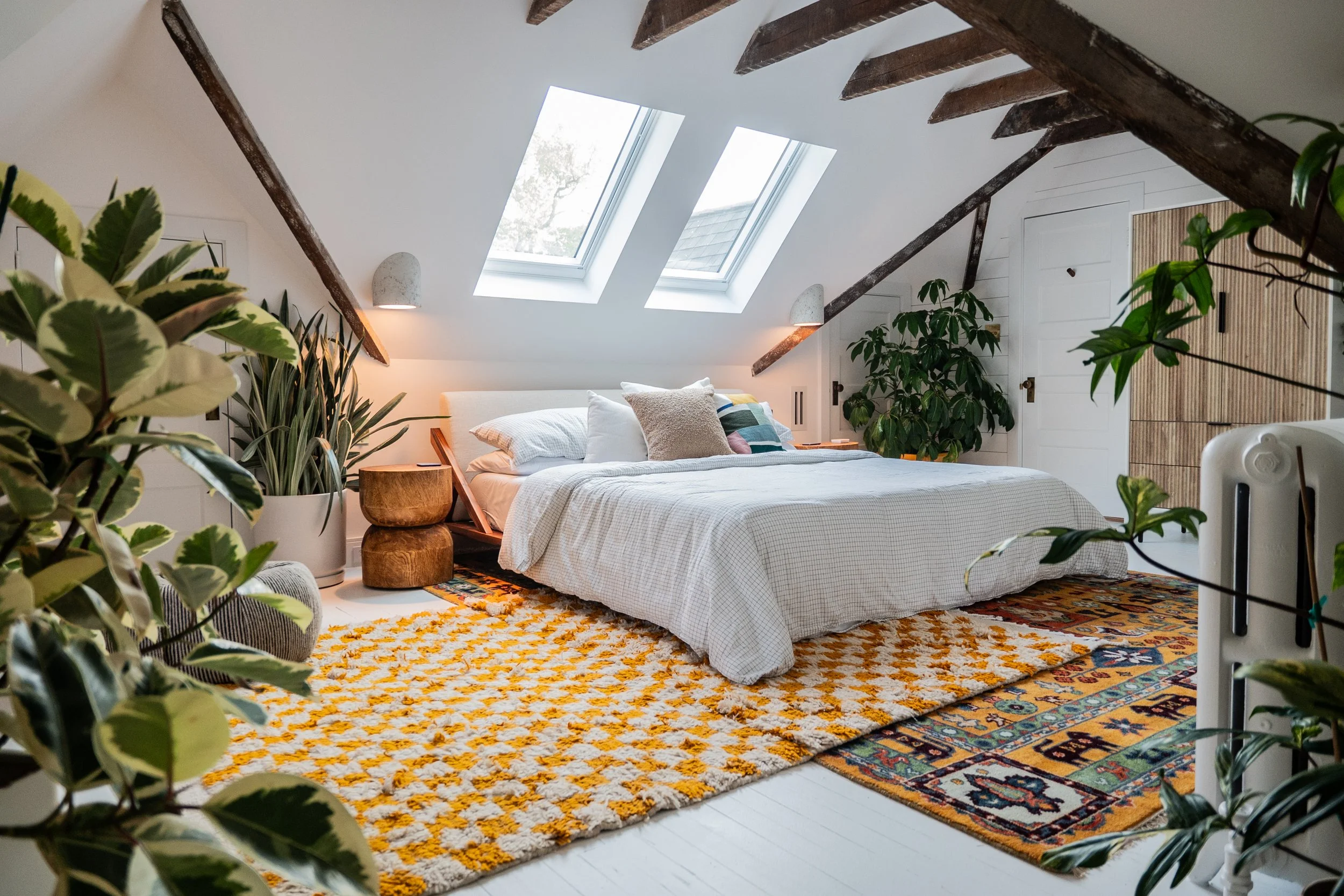 Cozy bedroom in an attic with sloped ceiling, solar skylights, a large bed with white bedding, side tables, multiple plants, and colorful patterned rugs.