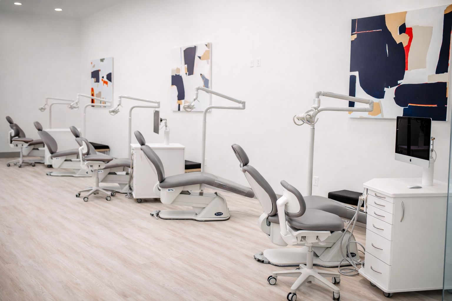 A row of dental or medical chairs with overhead lights and dental equipment in a clean, modern clinic with white walls and abstract artwork on the wall.