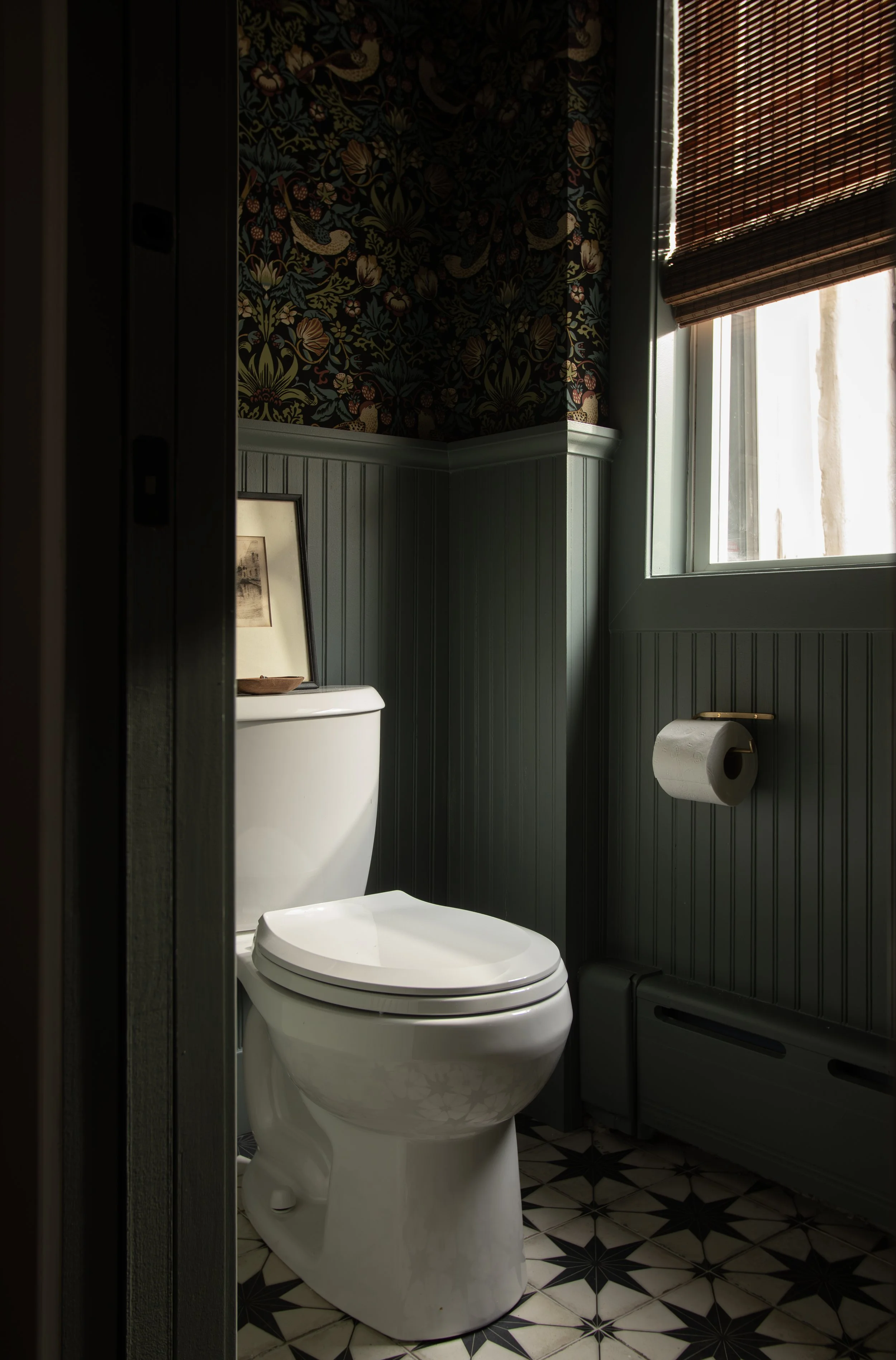 A small bathroom with dark green wainscoting, floral wallpaper, a white toilet with a framed picture and a bowl on top, a window with bamboo blinds, and a black and white patterned tile floor.
