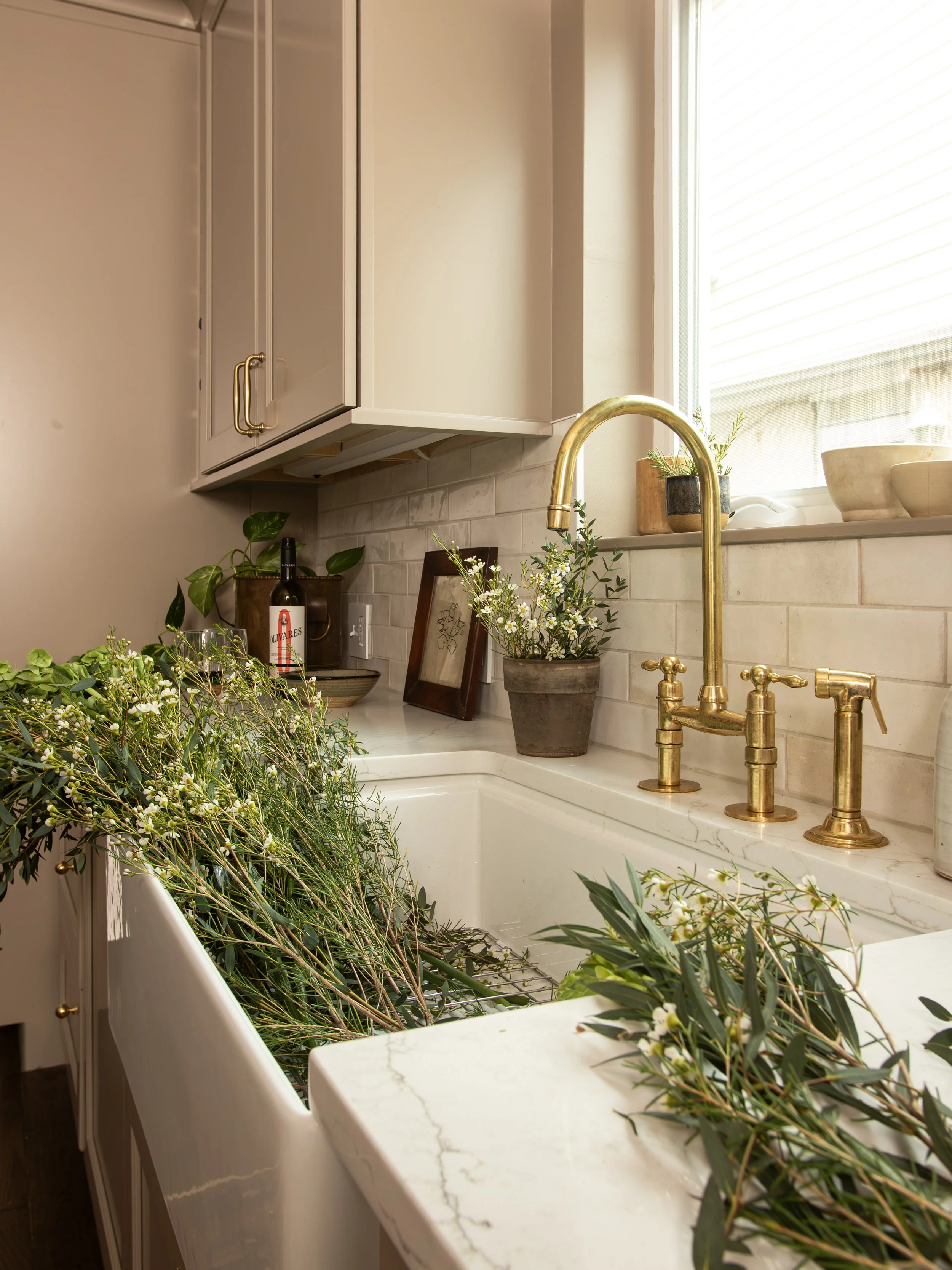 A kitchen sink with a gold faucet, filled with green branches and small white flowers, next to a window with potted plants, a framed picture, and decorative bowls on the windowsill.