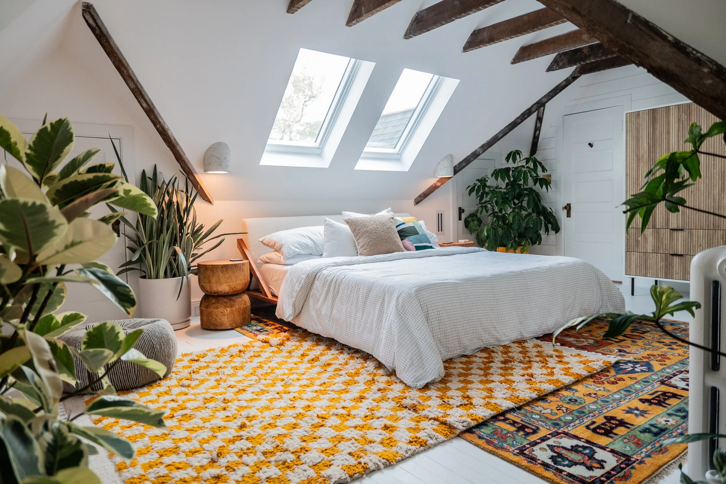 A bright bedroom with a sloped ceiling and skylights. It features a white bed with multiple pillows, a patterned yellow and orange rug, and various green plants. There are wooden beams and a small door on the wall.