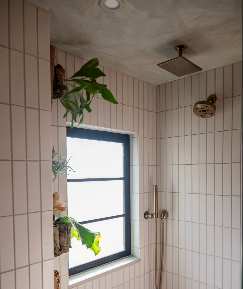Shower area with beige tiled walls, a window, a rainfall showerhead on the ceiling, a handheld shower, and hanging plants near the window.