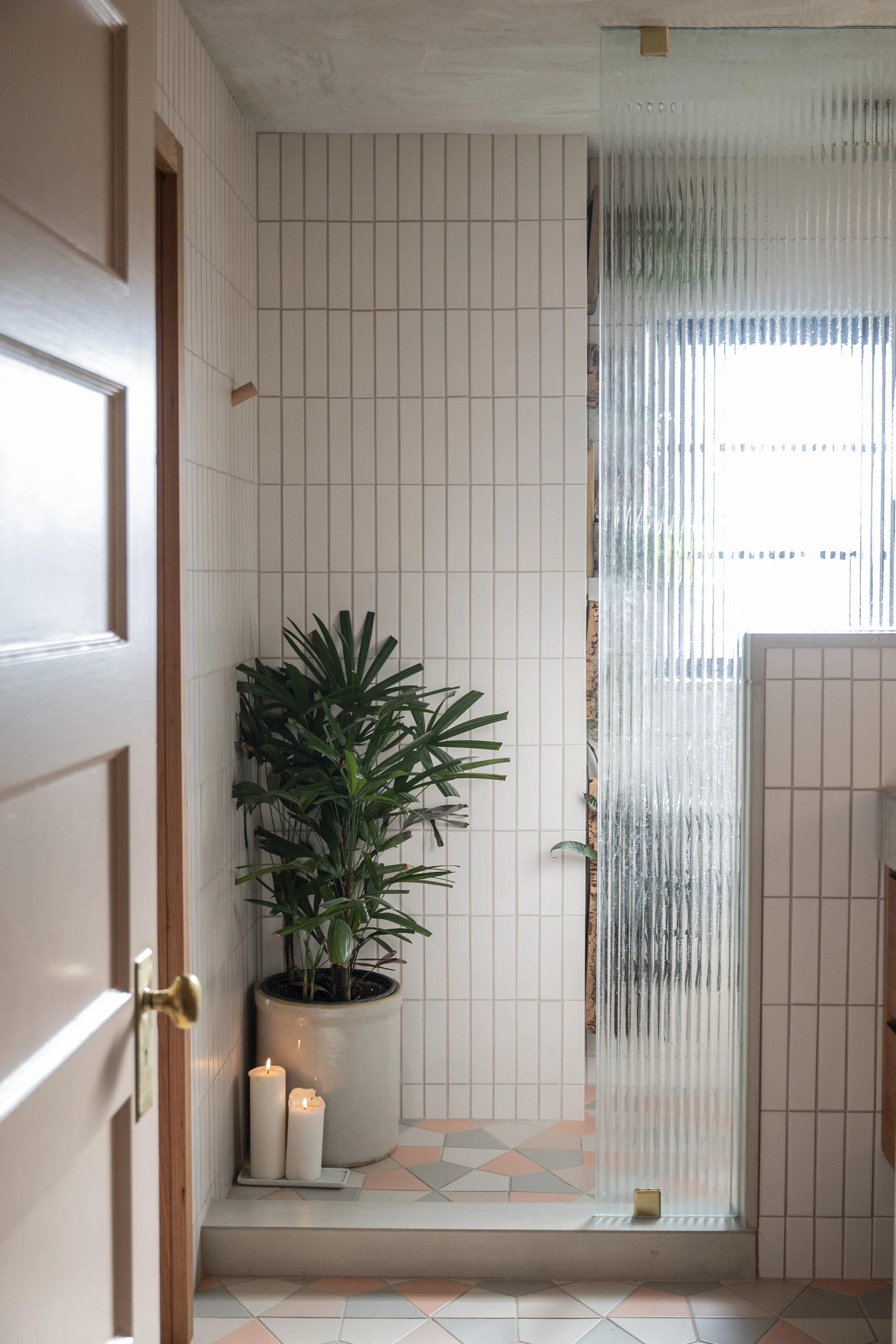 A small bathroom with a large potted green plant and three white candles beside it. The bathroom has white tiled walls, a mosaic tile floor with pink, gray, and white colors, and a partial glass shower door. Natural light is coming through a window.