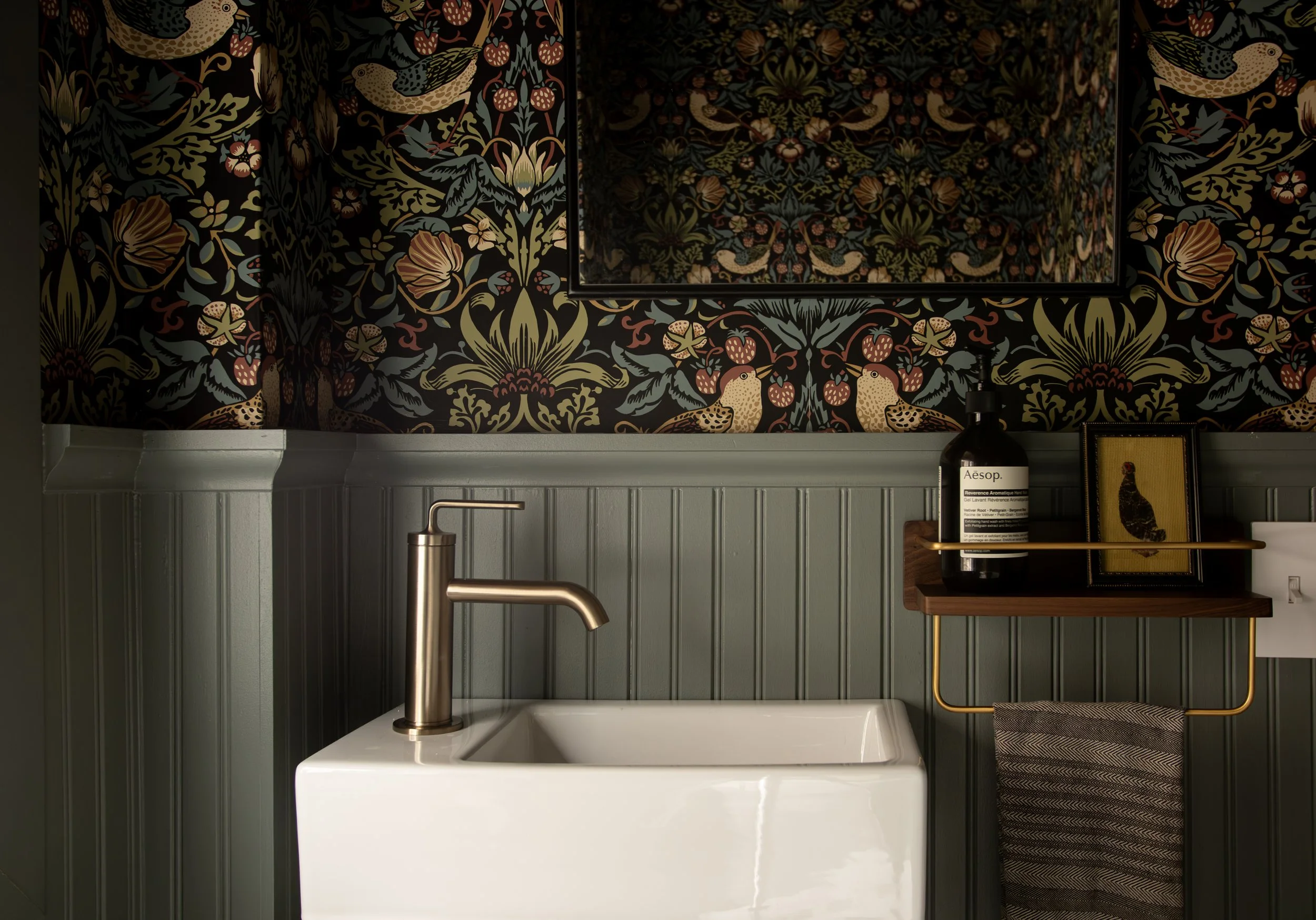 A bathroom with leaf-patterned dark wallpaper featuring birds and flowers, a white rectangular sink with a brushed bronze faucet, and a wooden and brass shelf holding a black bottle, framed bird artwork, and a pilot-style towel hanger with a patterne