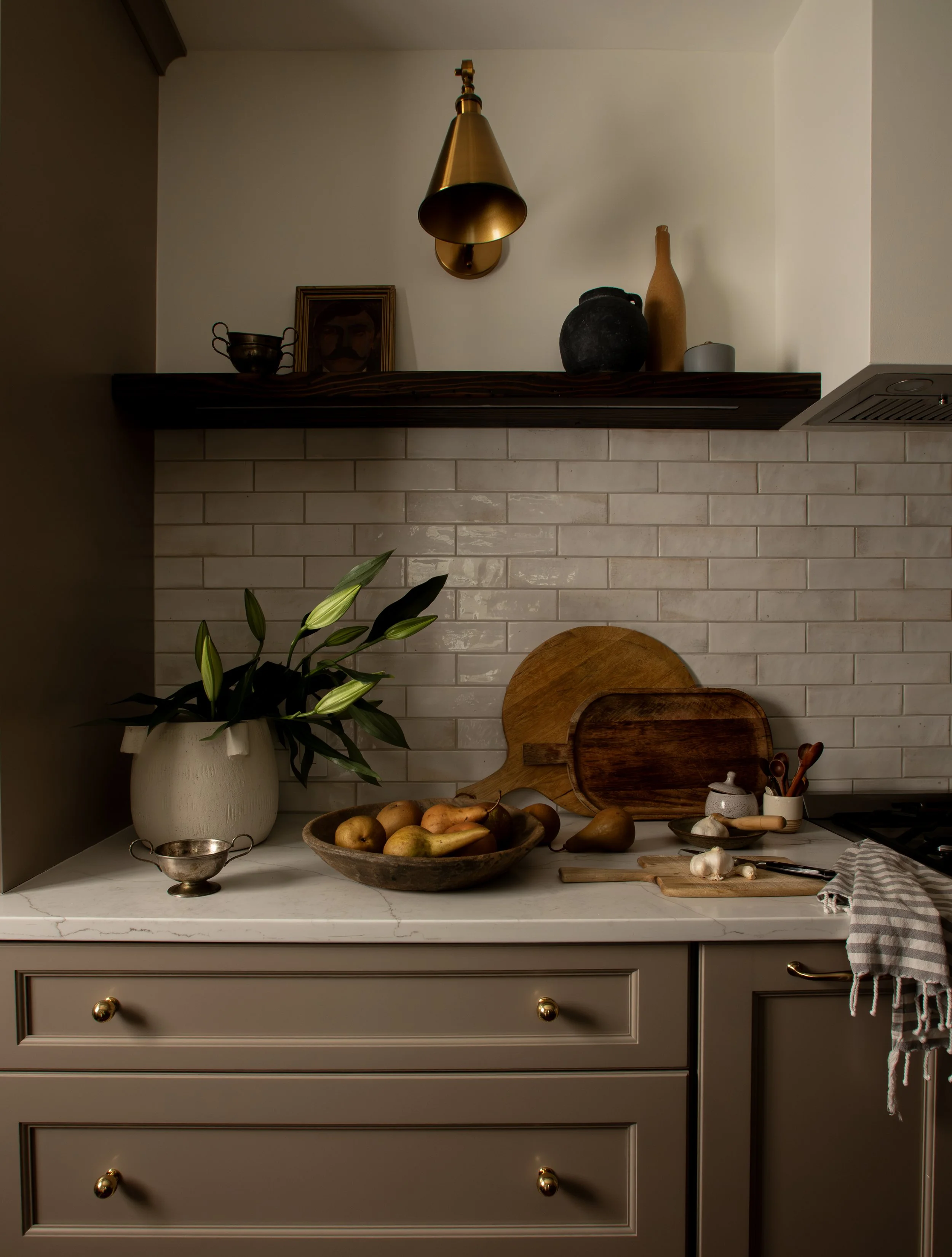 A kitchen counter with a potted plant, bowls of potatoes, garlic, wooden cutting boards, and kitchen utensils, with a beige drawer cabinet and a white tiled backsplash.