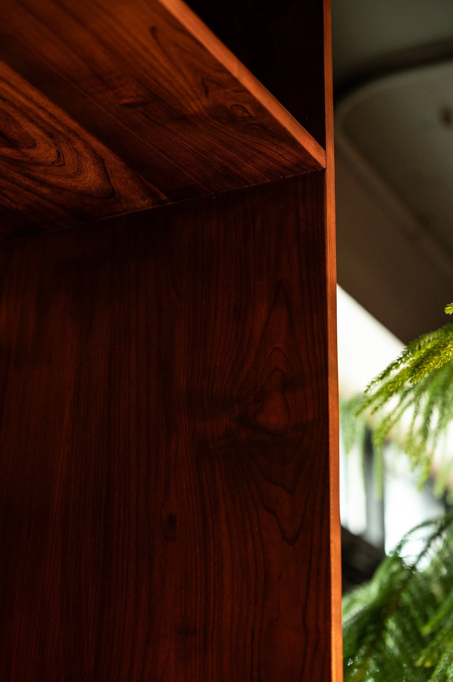 Close-up of a wooden piece of furniture, with a visible wood grain pattern, set against a background of blurred green foliage and part of a ceiling.