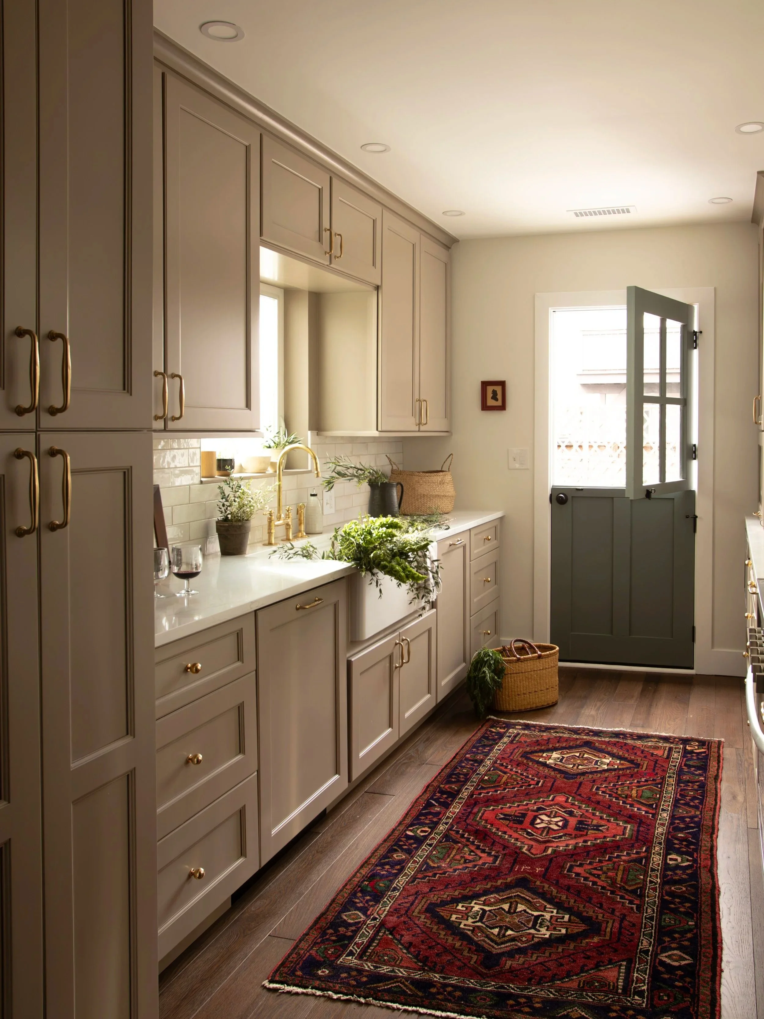 A cozy kitchen with beige cabinets, a white countertop, a gold faucet, and a small window. There are wine glasses, plants, a woven basket, and a colorful patterned rug on the wooden floor.