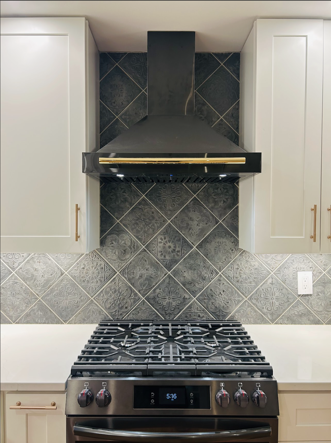 A kitchen stove with a black range hood above it, surrounded by white cabinetry, and decorative gray tile backsplash with intricate patterns.