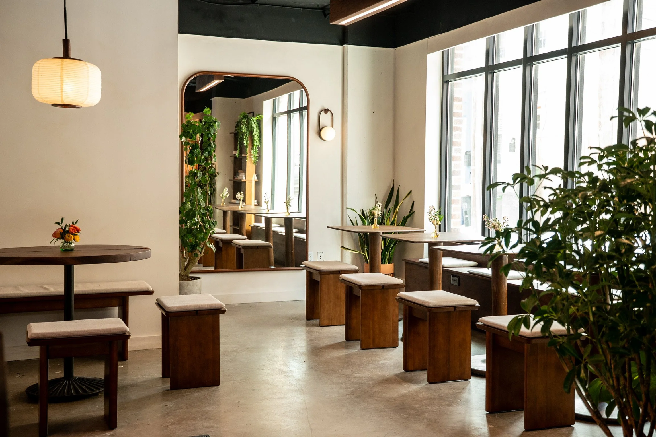 Interior of a modern cafe or restaurant with wooden tables, small vases with flowers, and large windows allowing natural light. Green plants and a mirror decorate the space, creating a cozy ambiance.