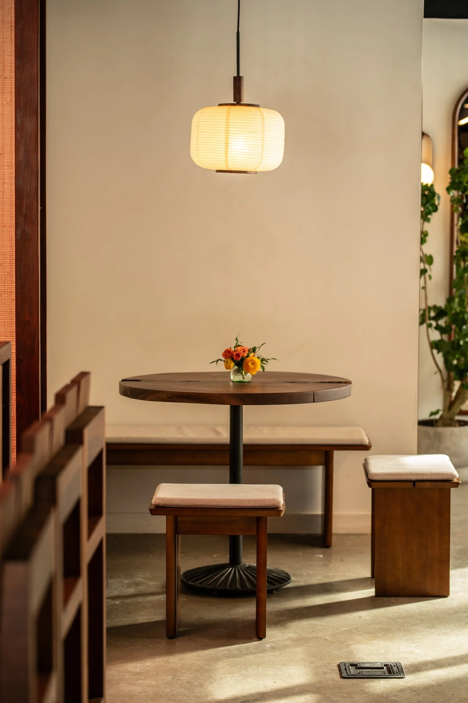 Interior of a restaurant or cafe featuring a round wooden table with a small flower arrangement, wooden stools with cushioned seats, a hanging paper lantern light, and a beige wall with some greenery on the right side.