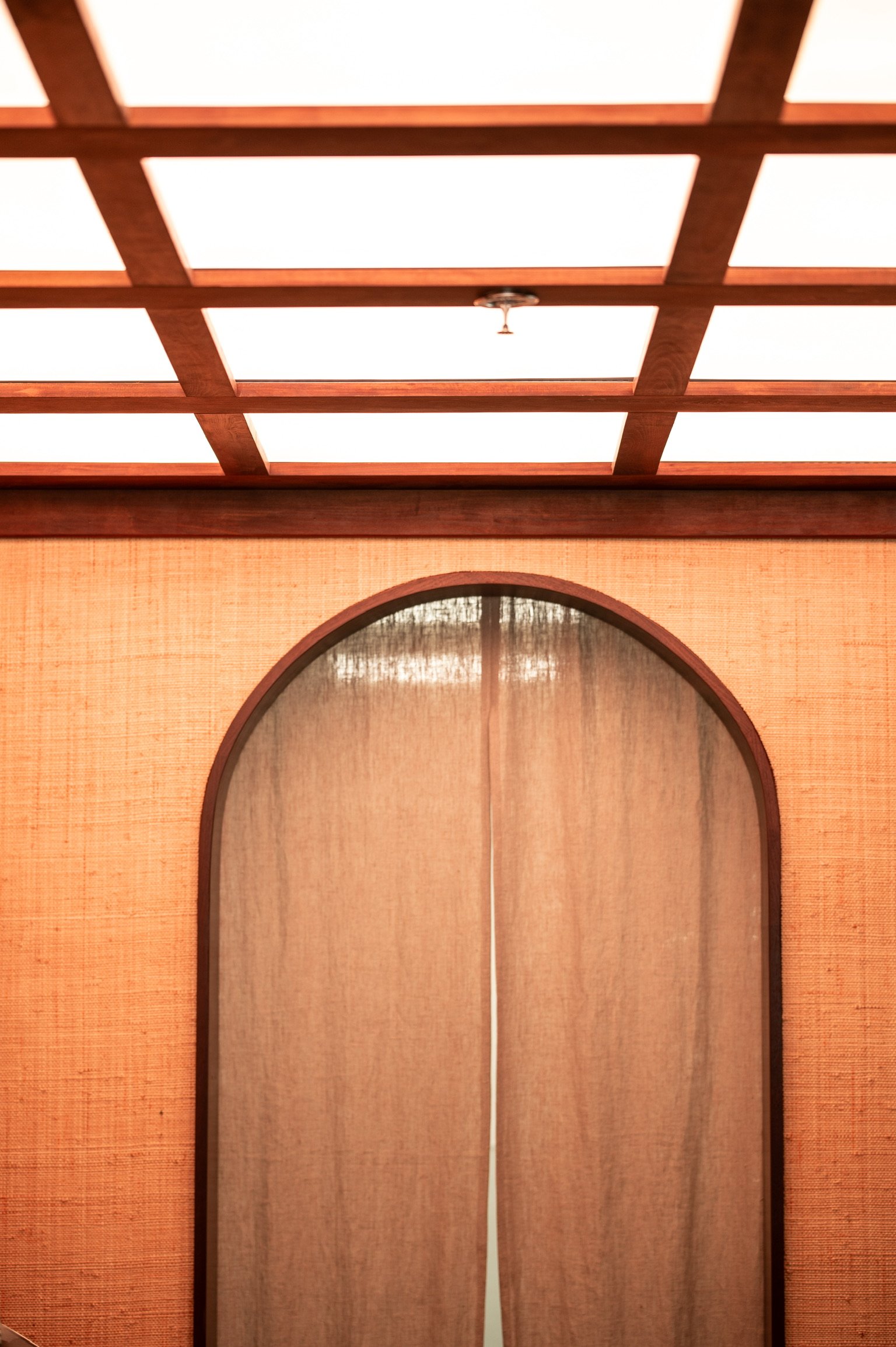 View of an indoor space featuring a wooden lattice ceiling and arched window with a curtain.
