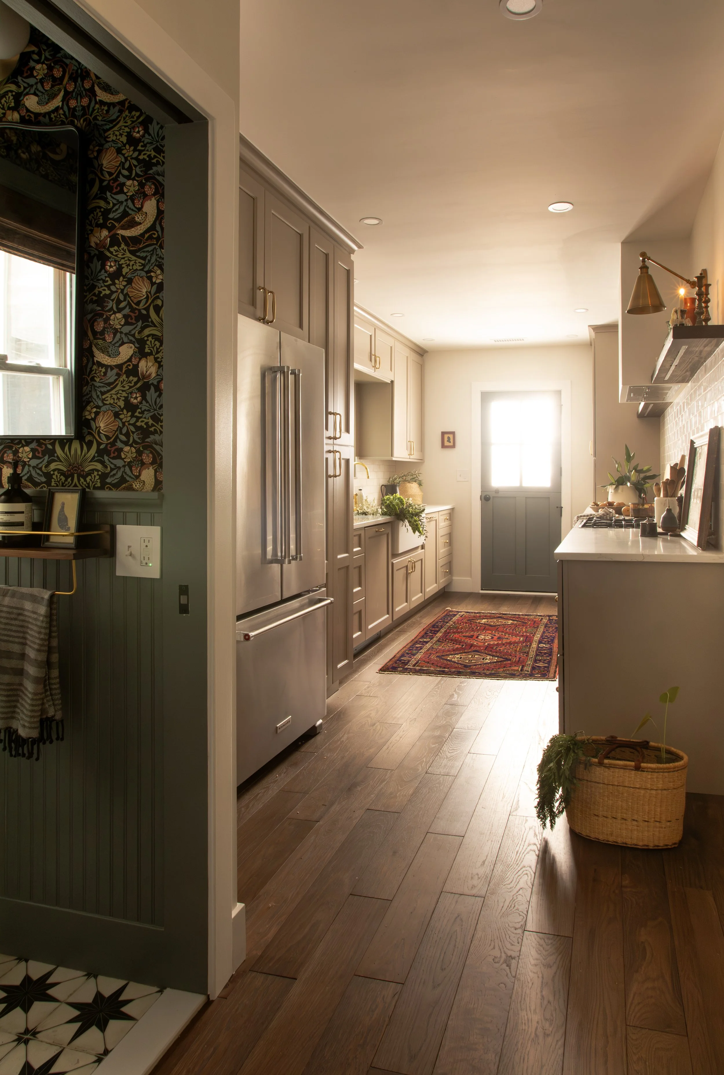 A cozy, well-lit kitchen with beige cabinetry, stainless steel refrigerator, dark hardwood floors, a colorful patterned rug, and potted plants by the door.
